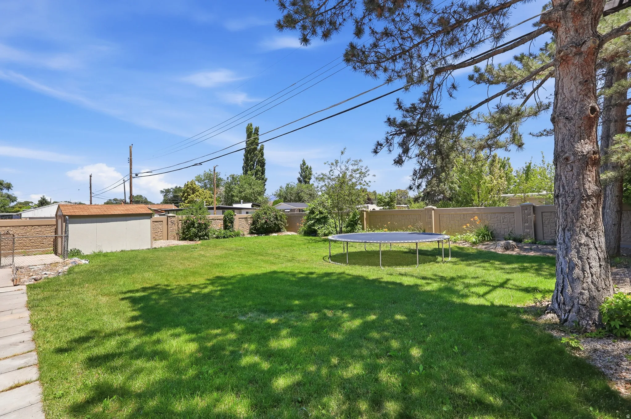 Expansive grassy backyard featuring a paved walkway, mature trees, a storage shed with a shingle roof, and a perimeter wall