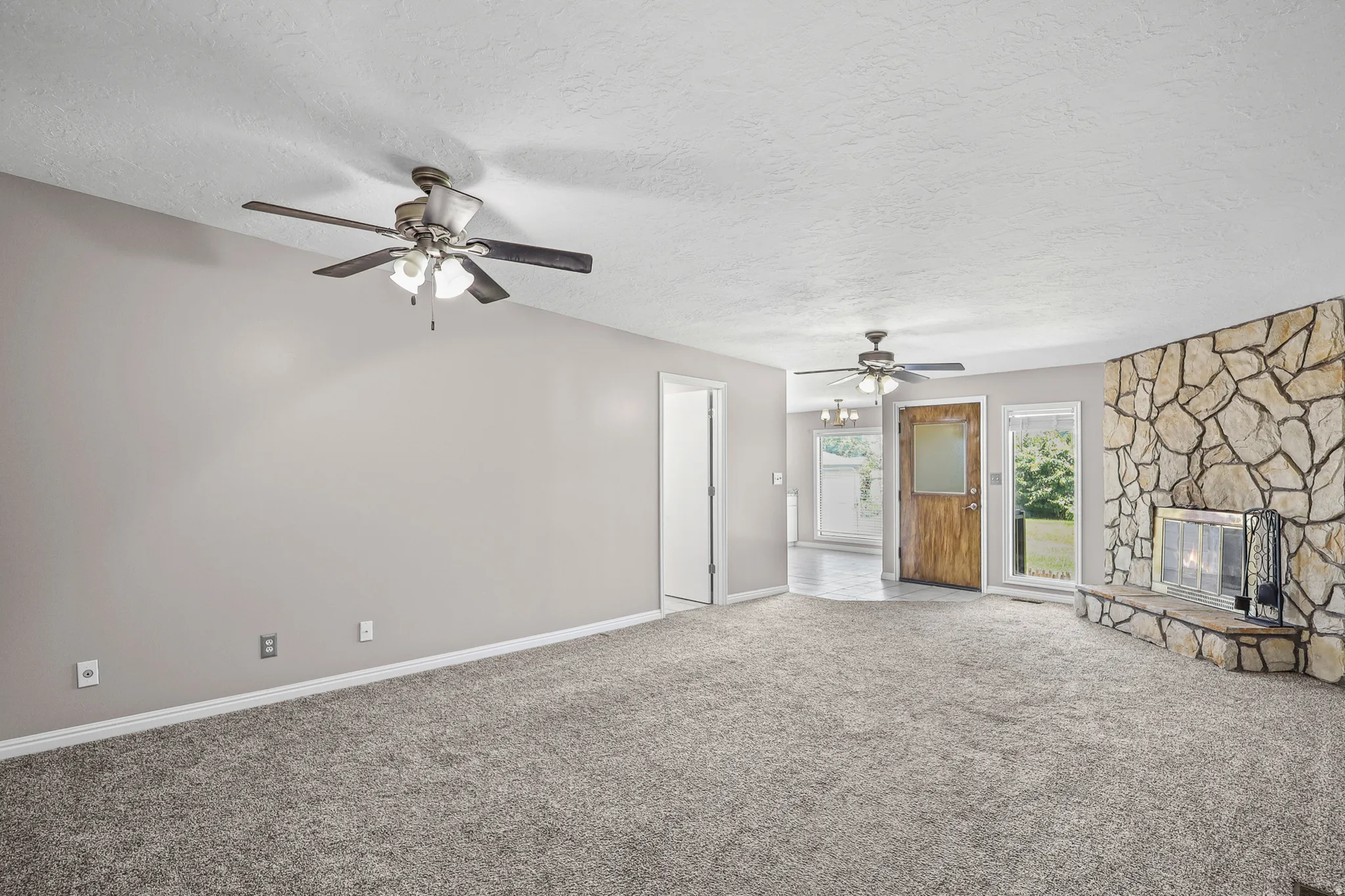 Spacious living area featuring neutral-toned walls, textured gray carpeting, and two ceiling fans with integrated lighting