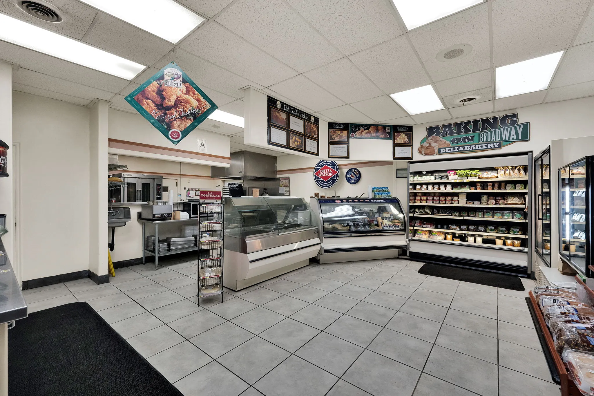 Commercial interior featuring light-toned square tile flooring, drop ceiling with integrated lighting, a service counter, multiple refrigerated display cases, and a rear service area with stainless steel fixtures