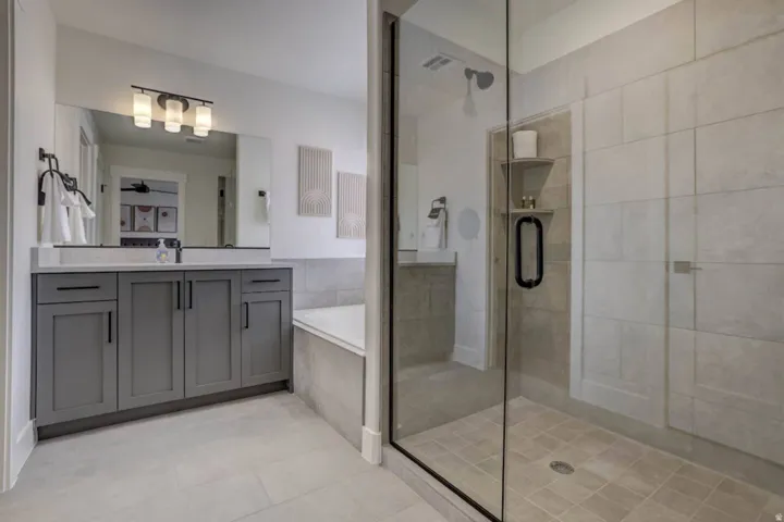 Glass-enclosed shower with tiled walls and built-in shelving, accompanied by a rectangular soaking tub and a dual-sink vanity with gray cabinetry and a light-toned countertop
