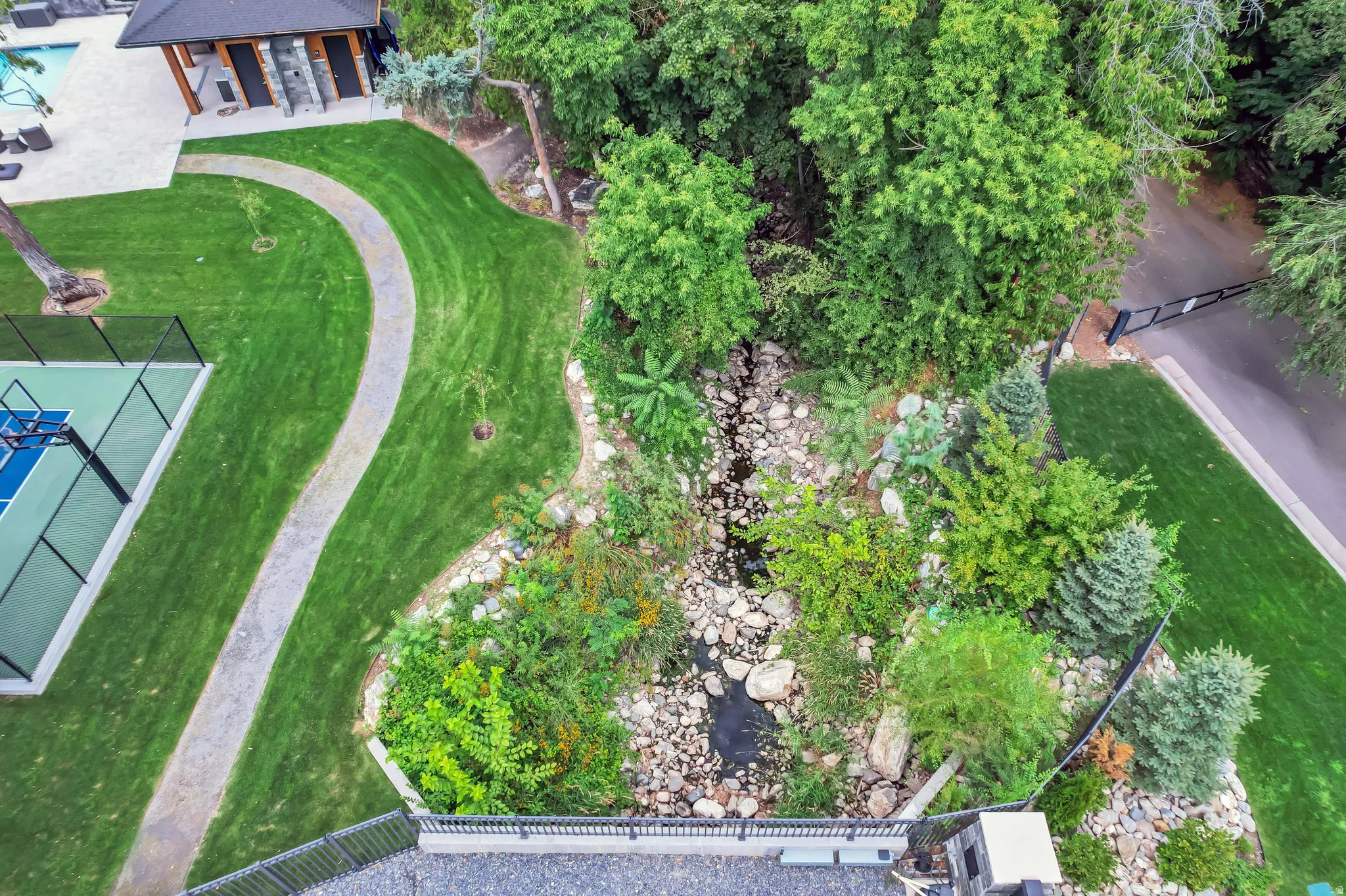 Expansive outdoor recreation area featuring a full-size sports court, a meandering paved pathway, a natural rock creek bed, a stone patio with an adjacent structure, and mature landscaping
