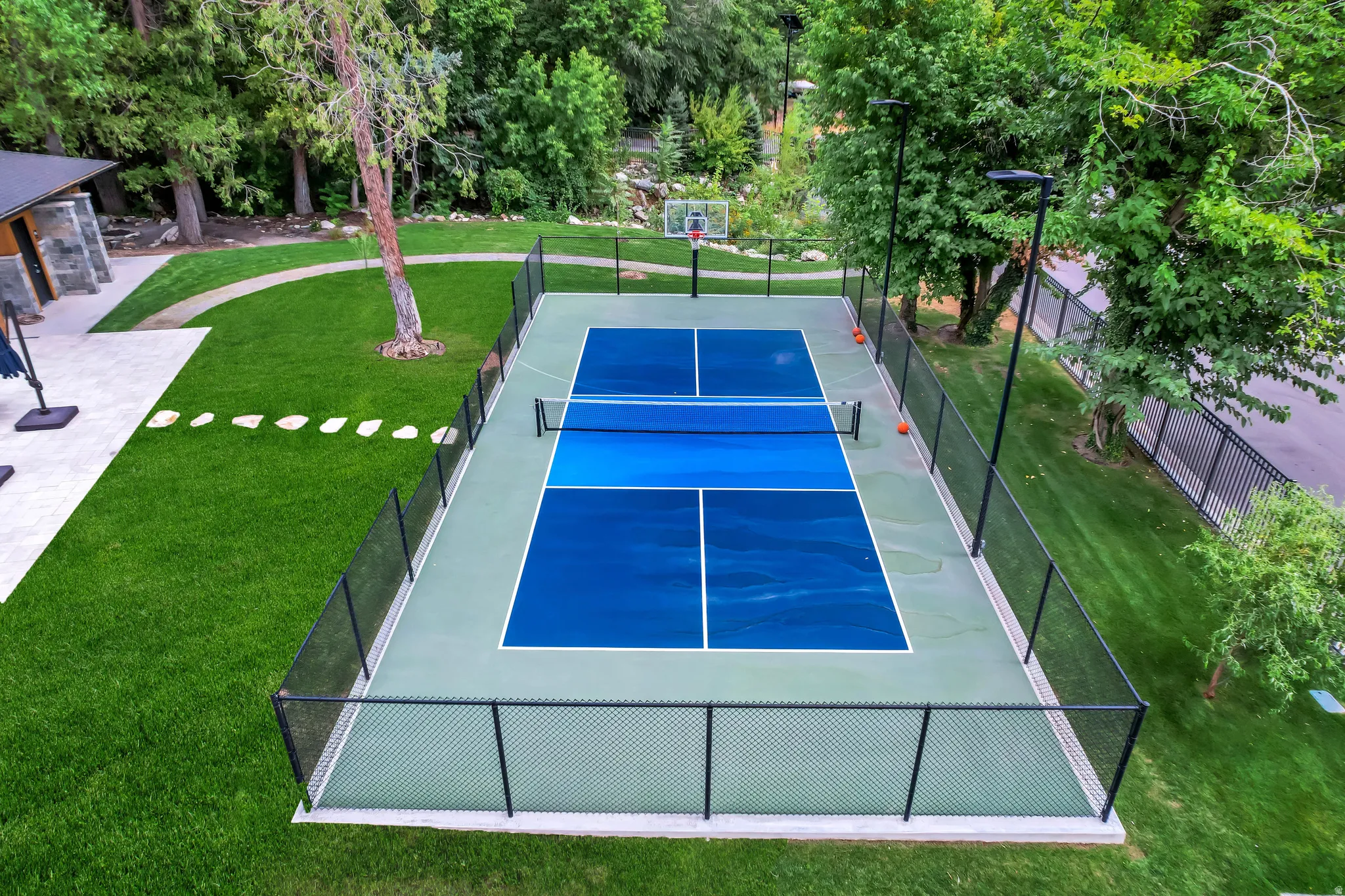 Multi-sport court with blue and green surfacing, featuring a basketball hoop and net, enclosed by a black chain-link fence