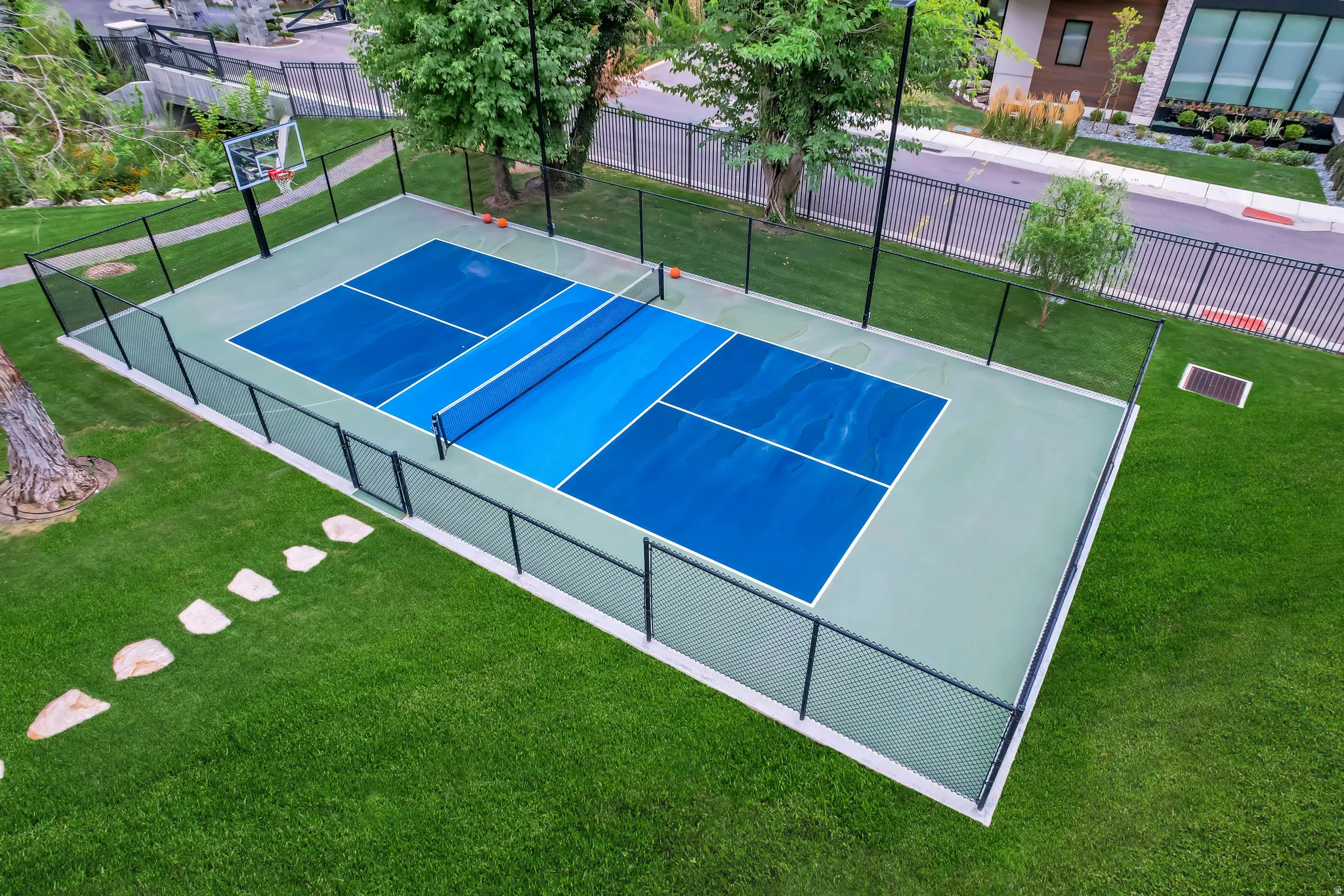 Fenced multi-sport court featuring a vibrant blue playing surface, light green surround, and a basketball hoop