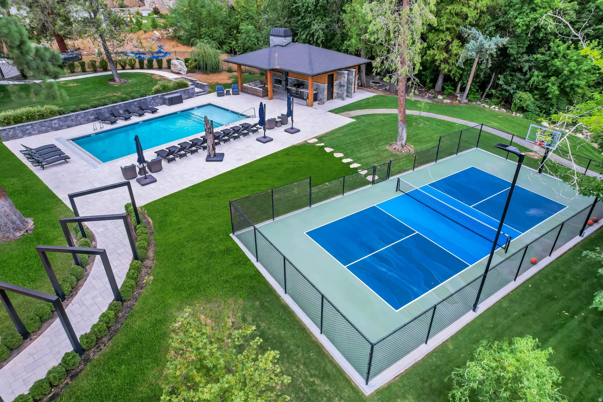 Expansive outdoor living area featuring a rectangular swimming pool with surrounding paver patio, a covered outdoor kitchen structure with stone accents, a multi-sport court with fencing and lighting, and a paved walkway with decorative archways