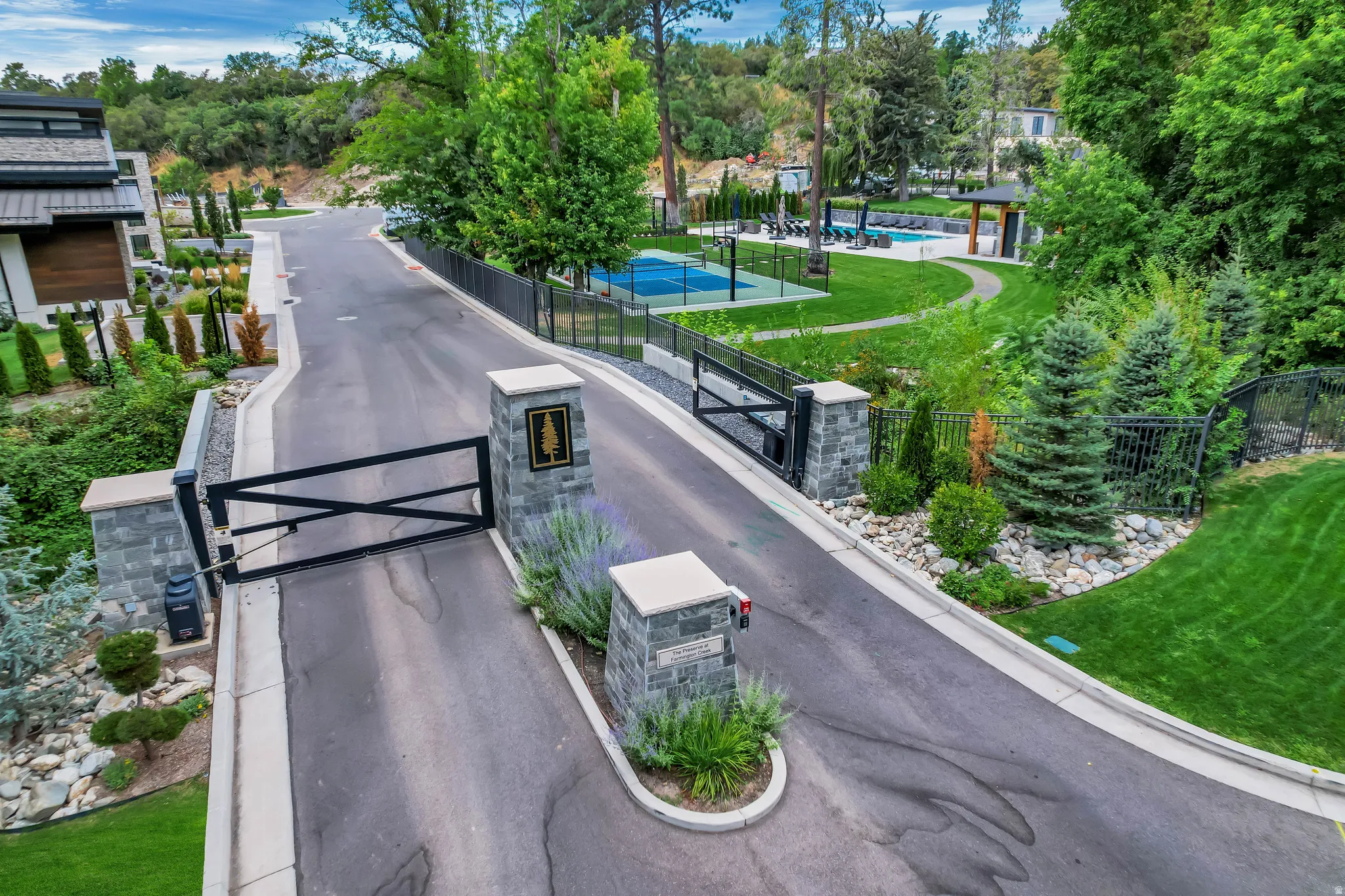 Gated community entrance featuring stone pillars, paved drive, mature landscaping, and proximity to community amenities including a pool and sports court
