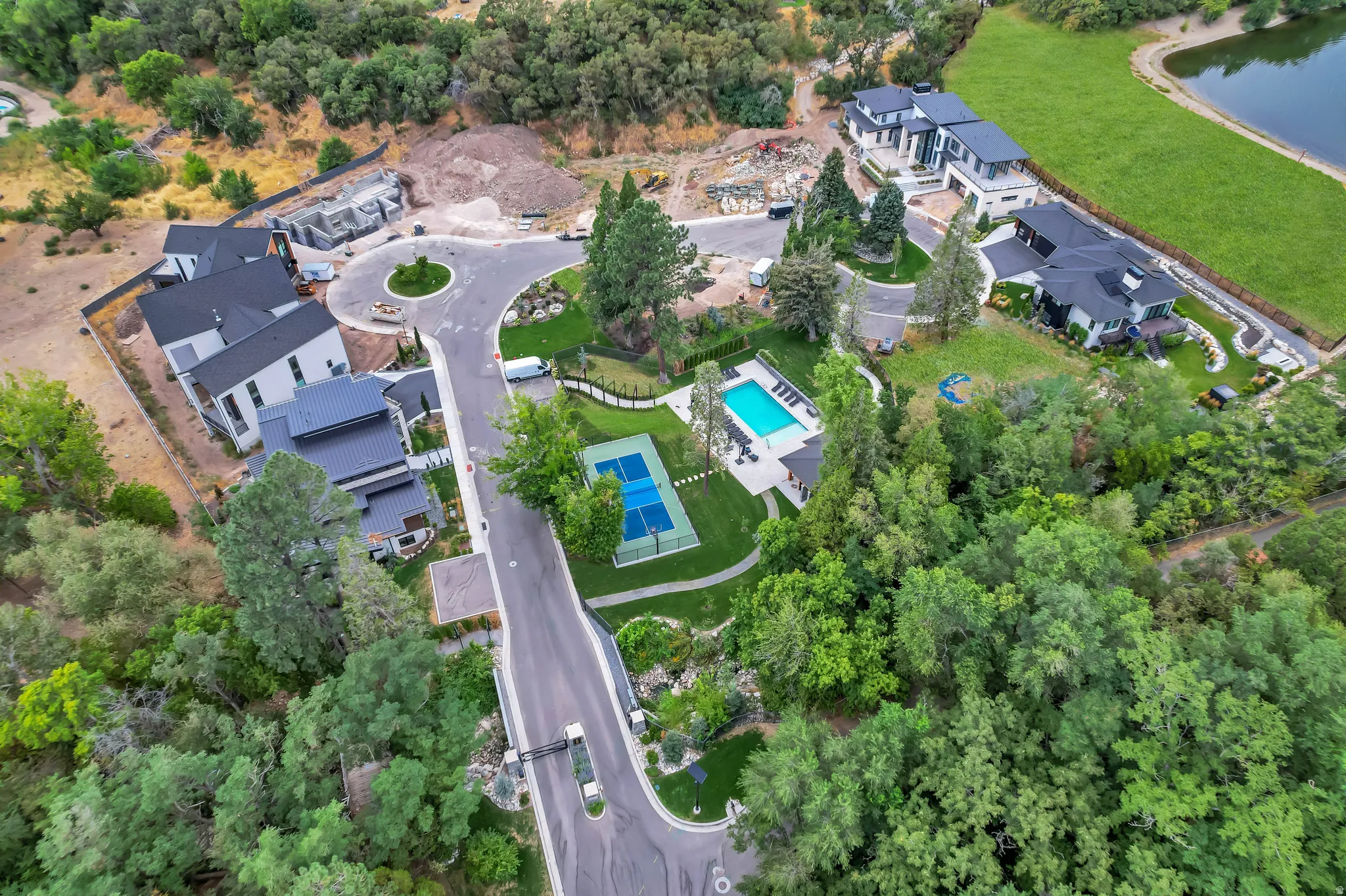 Aerial view of a residential community with contemporary architecture, featuring a private swimming pool, a dedicated tennis court, mature tree landscaping, and a curvilinear paved road