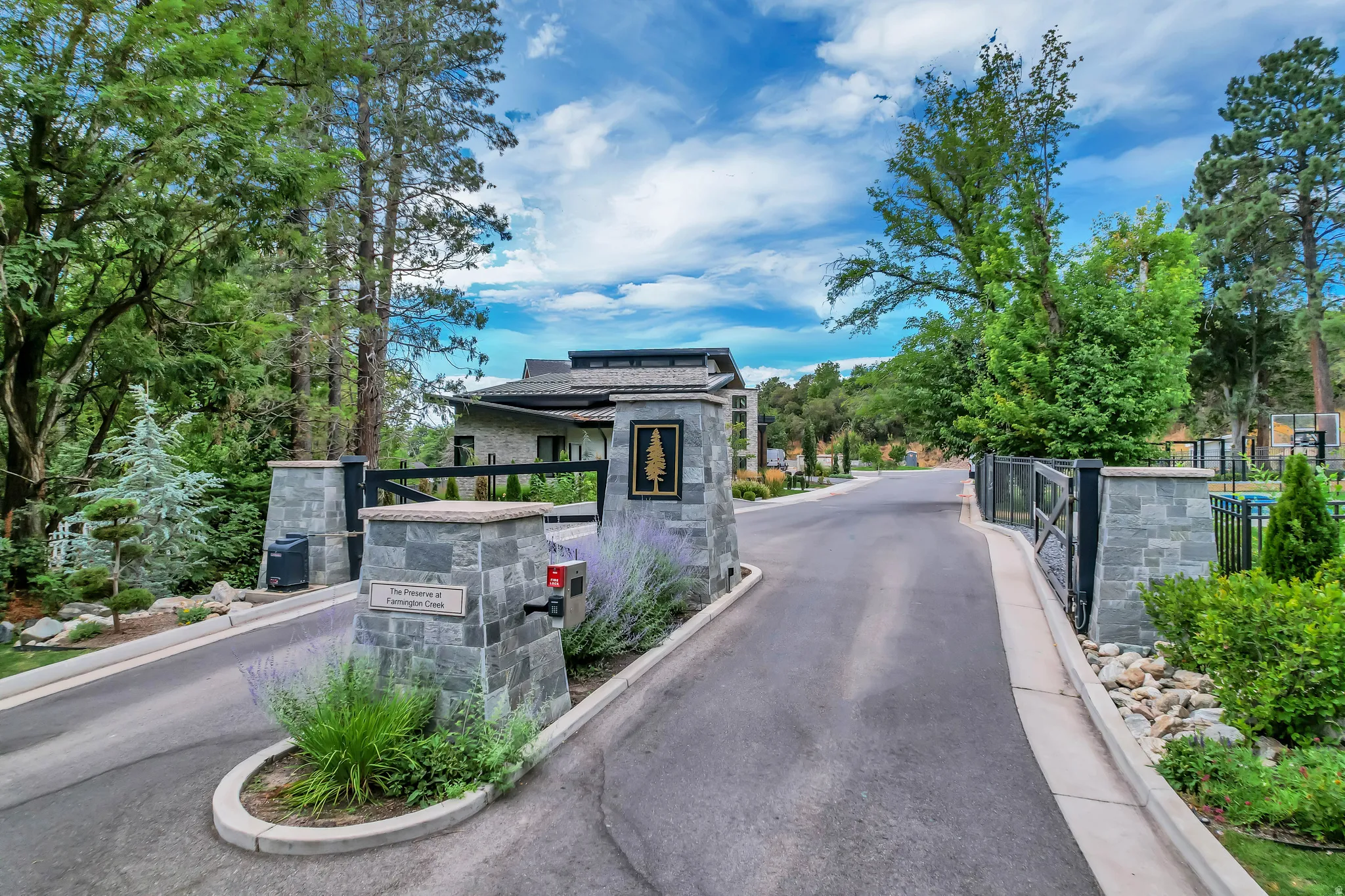 Gated property entrance featuring stone pillars, black metal fencing, and mature landscaping