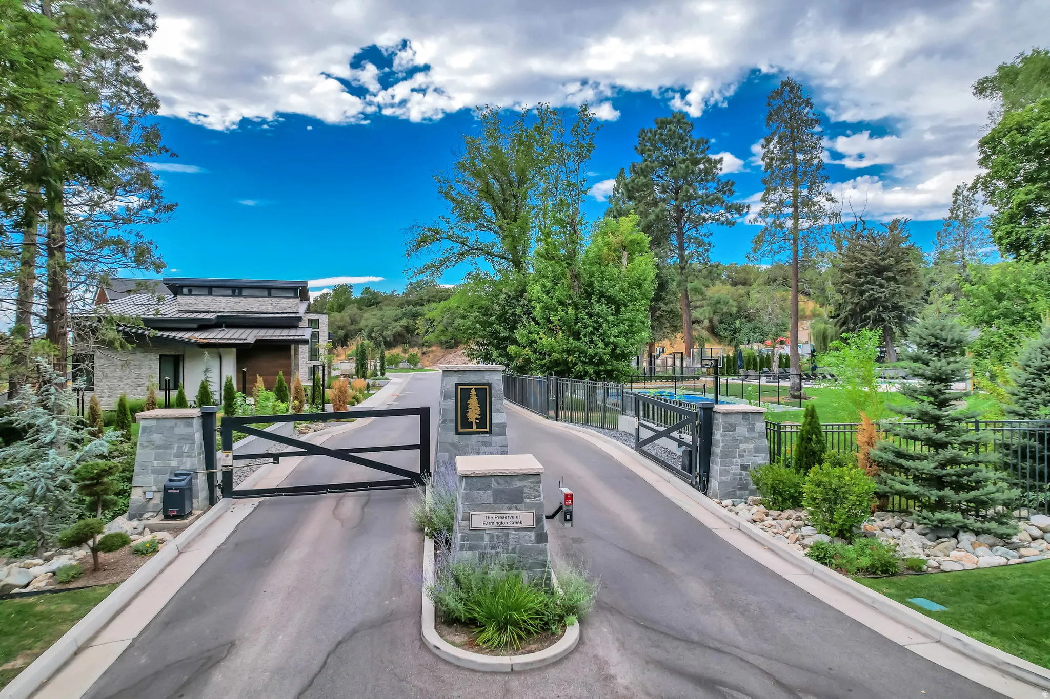 Gated entrance featuring stone pillars, black metal fencing, and a paved driveway, surrounded by mature trees and manicured landscaping