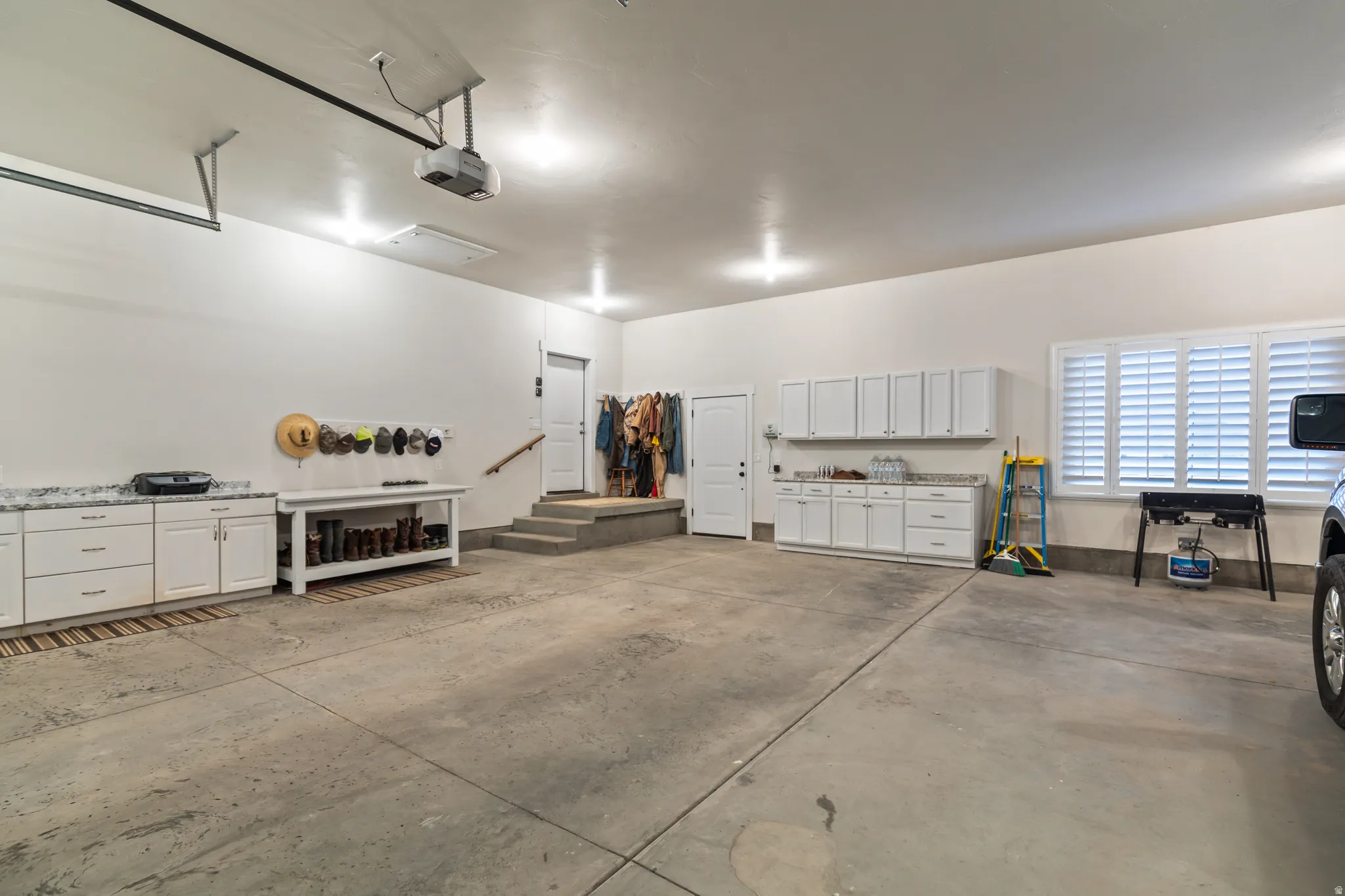 Spacious garage featuring white cabinetry with speckled countertops, multiple overhead light fixtures, and windows with white louvered shutters