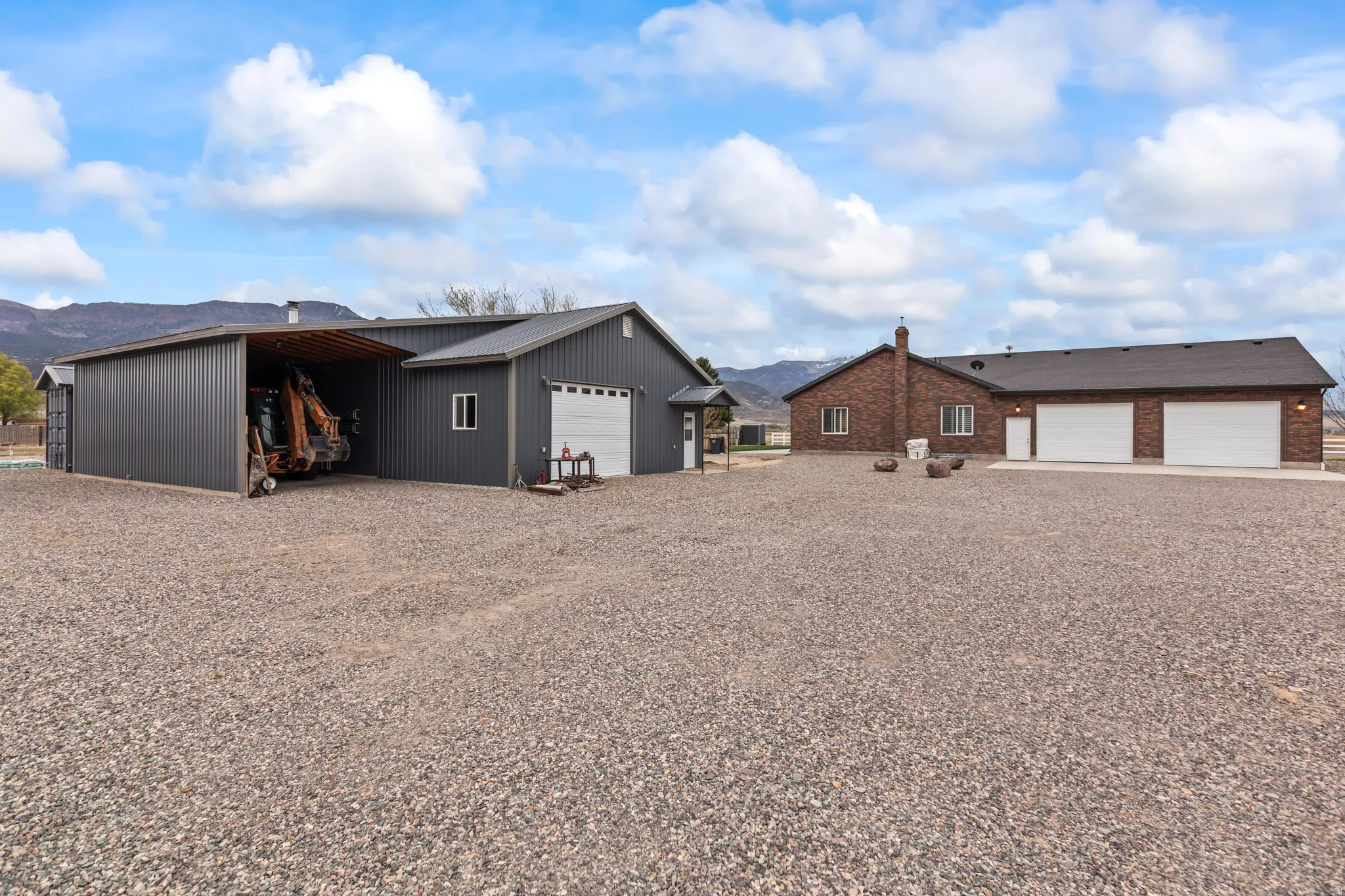 Gravel driveway presenting a brick-exterior residence with an attached three-car garage and a separate metal-sided outbuilding with an open-sided bay and an enclosed garage