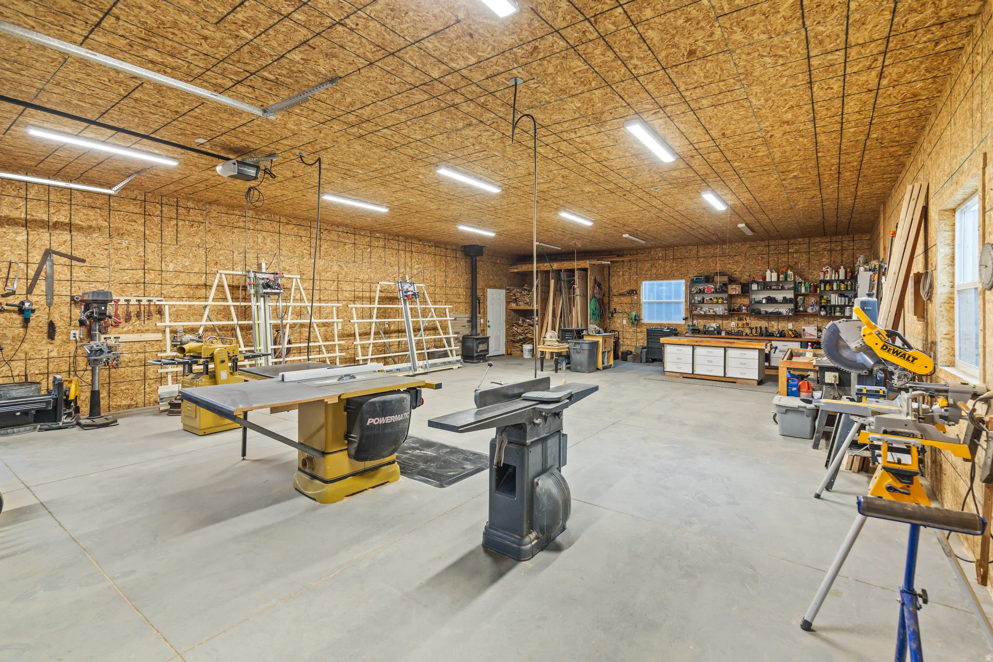 Spacious workshop featuring exposed plywood walls and ceiling, extensive overhead lighting, a concrete floor, and a built-in workbench with storage