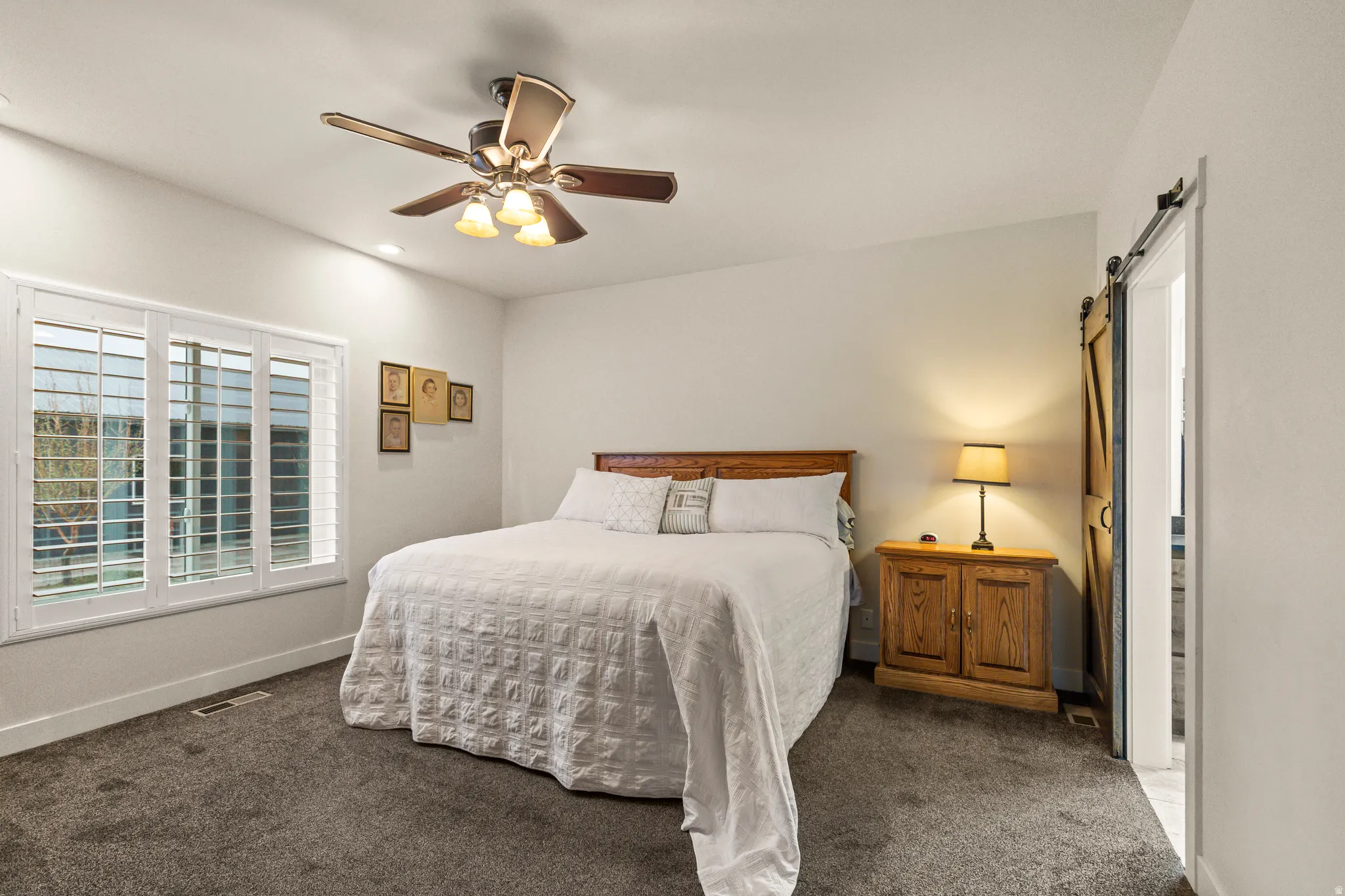 Carpeted bedroom featuring a ceiling fan with light fixture, triple window with white plantation shutters, a wood-finish barn door, and recessed lighting
