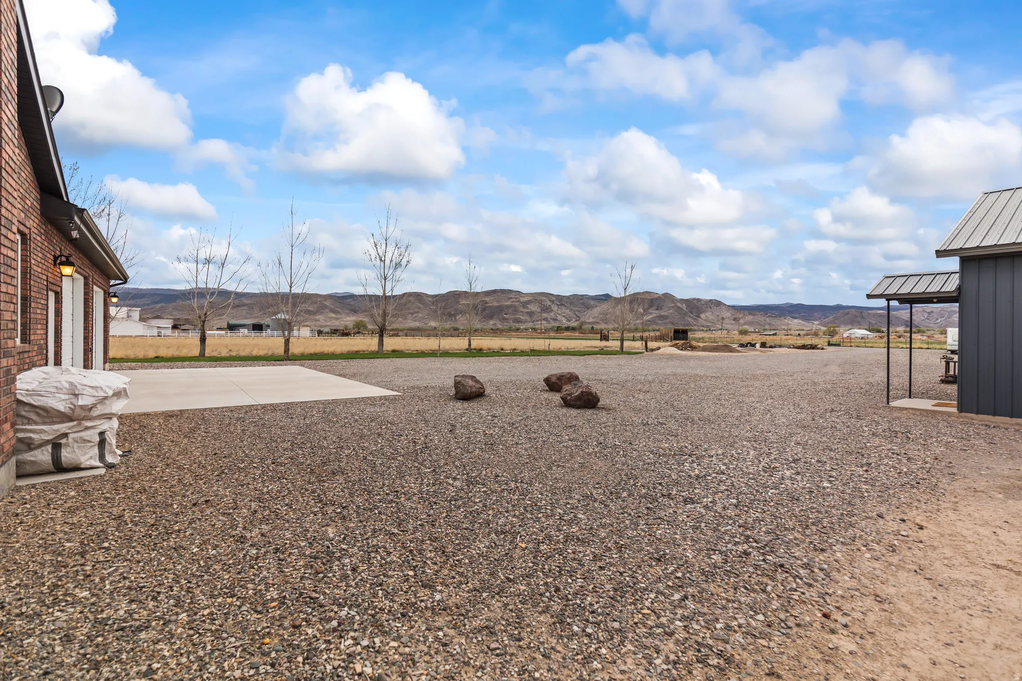 Expansive gravel lot with mountain backdrop, featuring a brick-clad structure with garage doors, a concrete pad, and a separate dark-paneled building with a covered entry