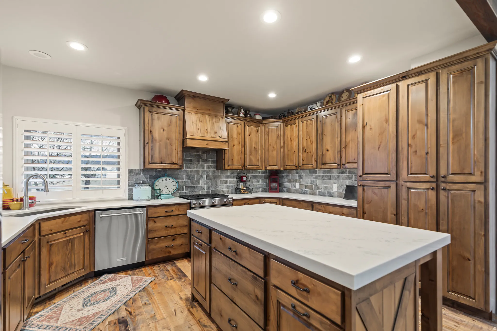 Kitchen featuring a large central island with a white countertop, extensive wood cabinetry, stainless steel dishwasher, a window with white shutters, and wood-finish flooring