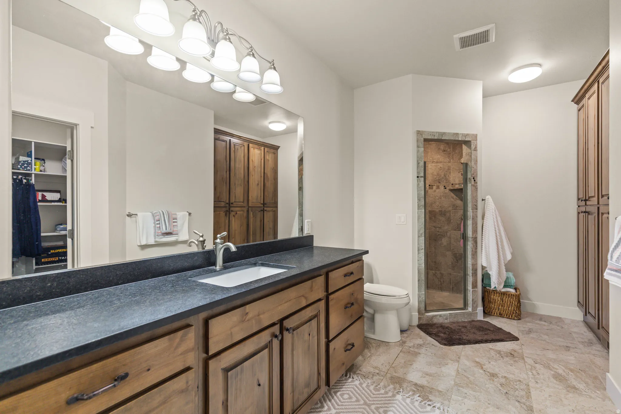 Spacious bathroom featuring a wood-finish vanity with a dark countertop and undermount sink, a tiled walk-in shower with a glass door, and extensive built-in cabinetry