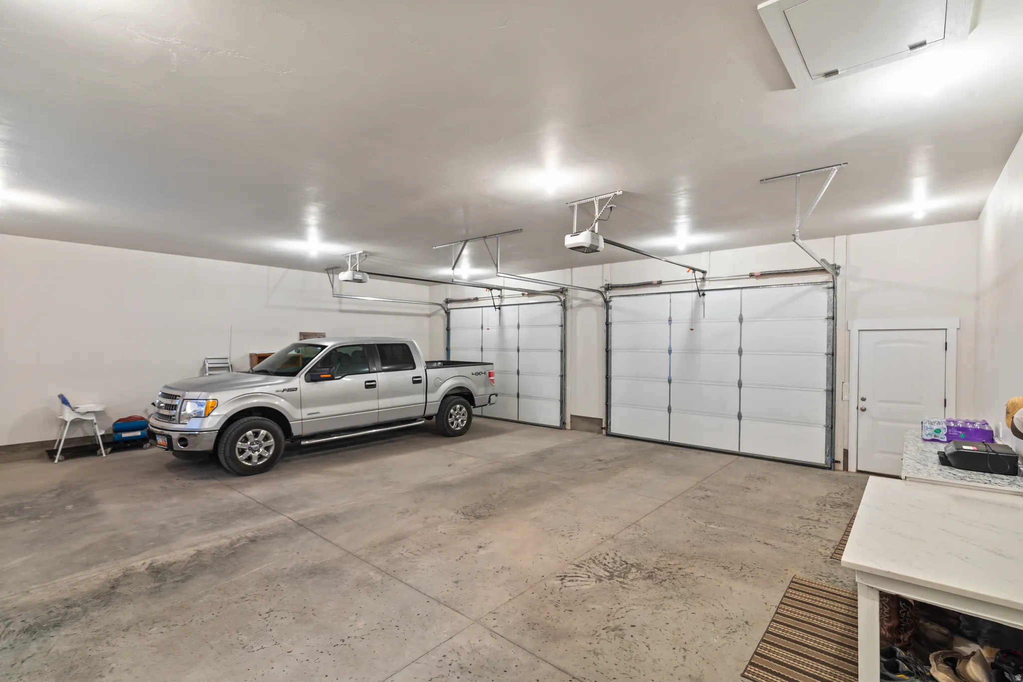 Spacious garage featuring two overhead doors, a pedestrian door, textured concrete flooring, and multiple ceiling-mounted light fixtures