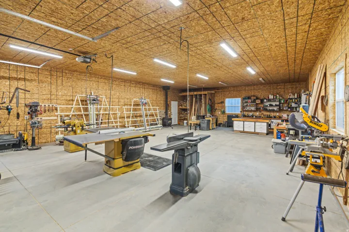 Spacious workshop featuring exposed plywood walls and ceiling, extensive overhead lighting, a concrete floor, and a built-in workbench with storage