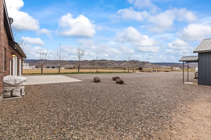 Expansive gravel lot with mountain backdrop, featuring a brick-clad structure with garage doors, a concrete pad, and a separate dark-paneled building with a covered entry