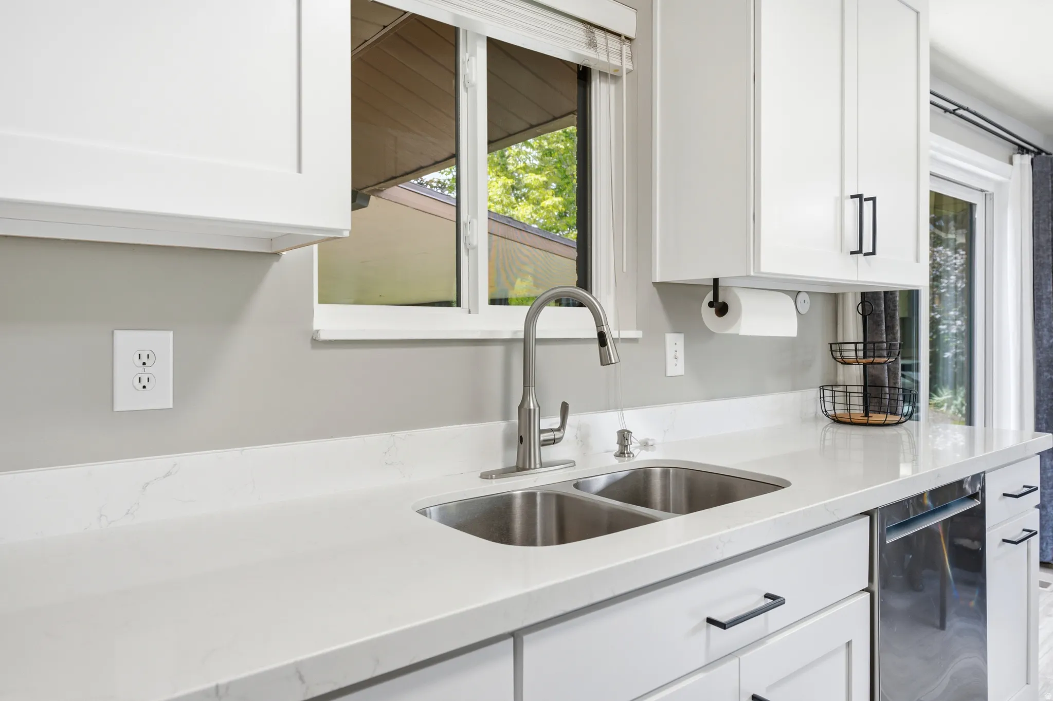Kitchen featuring a double stainless steel sink with a gooseneck faucet, light-toned countertop, white cabinetry with black hardware, and a built-in dishwasher
