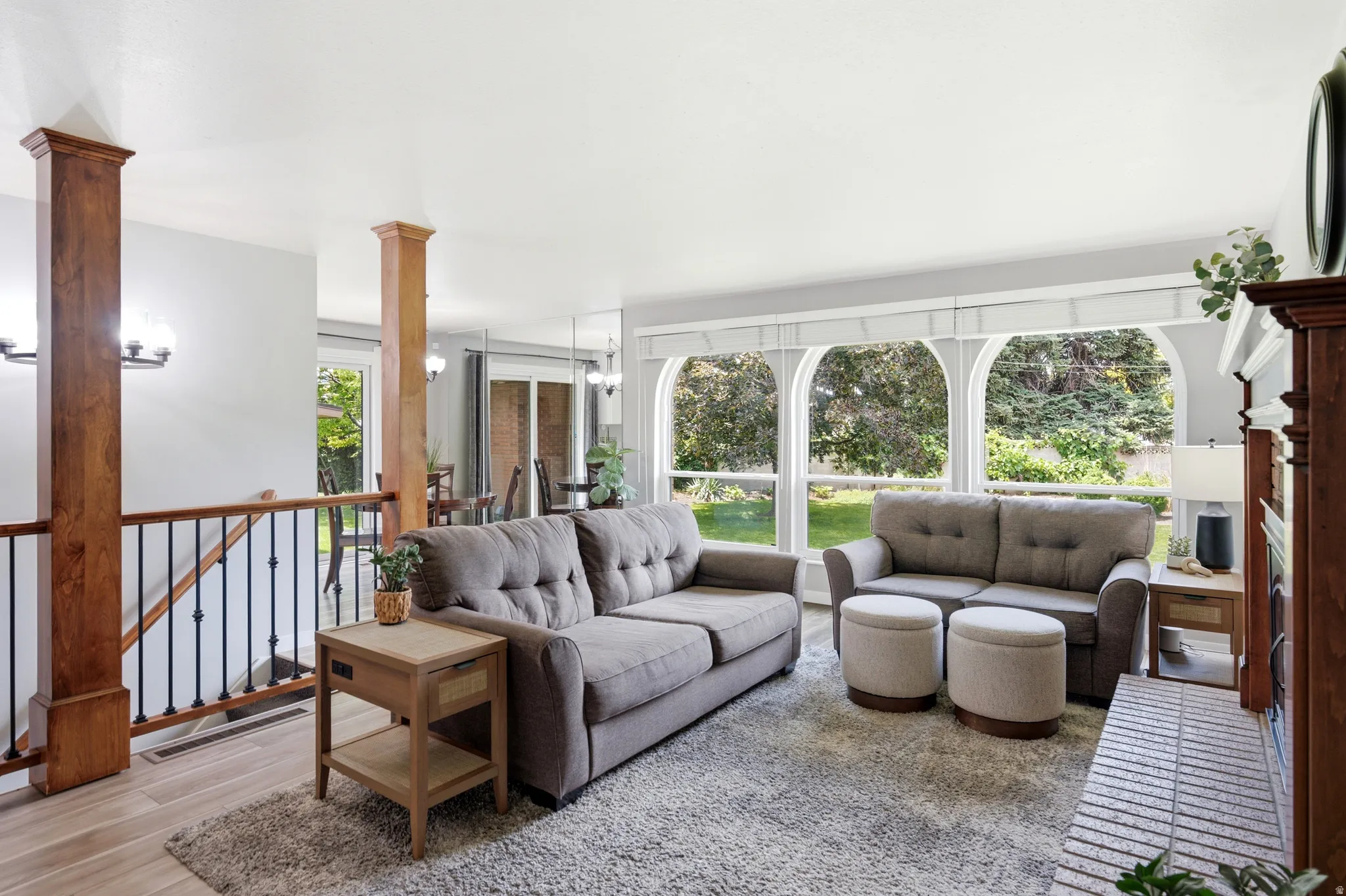 Spacious living area featuring large arched windows, light wood-finish flooring, a contemporary fireplace with a brick hearth, and a wooden staircase with wrought iron balusters