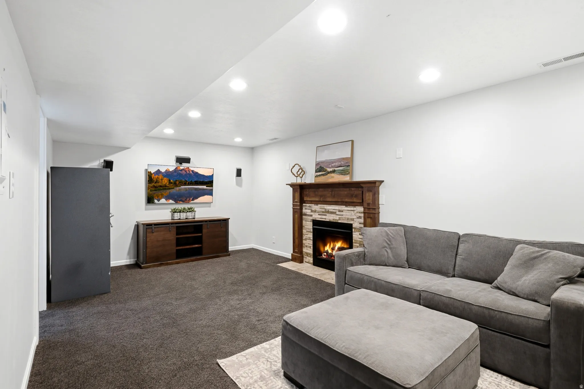 Recessed lighting illuminates this living space featuring dark carpet flooring, a stone-faced fireplace with a wood mantel, and built-in wall speakers