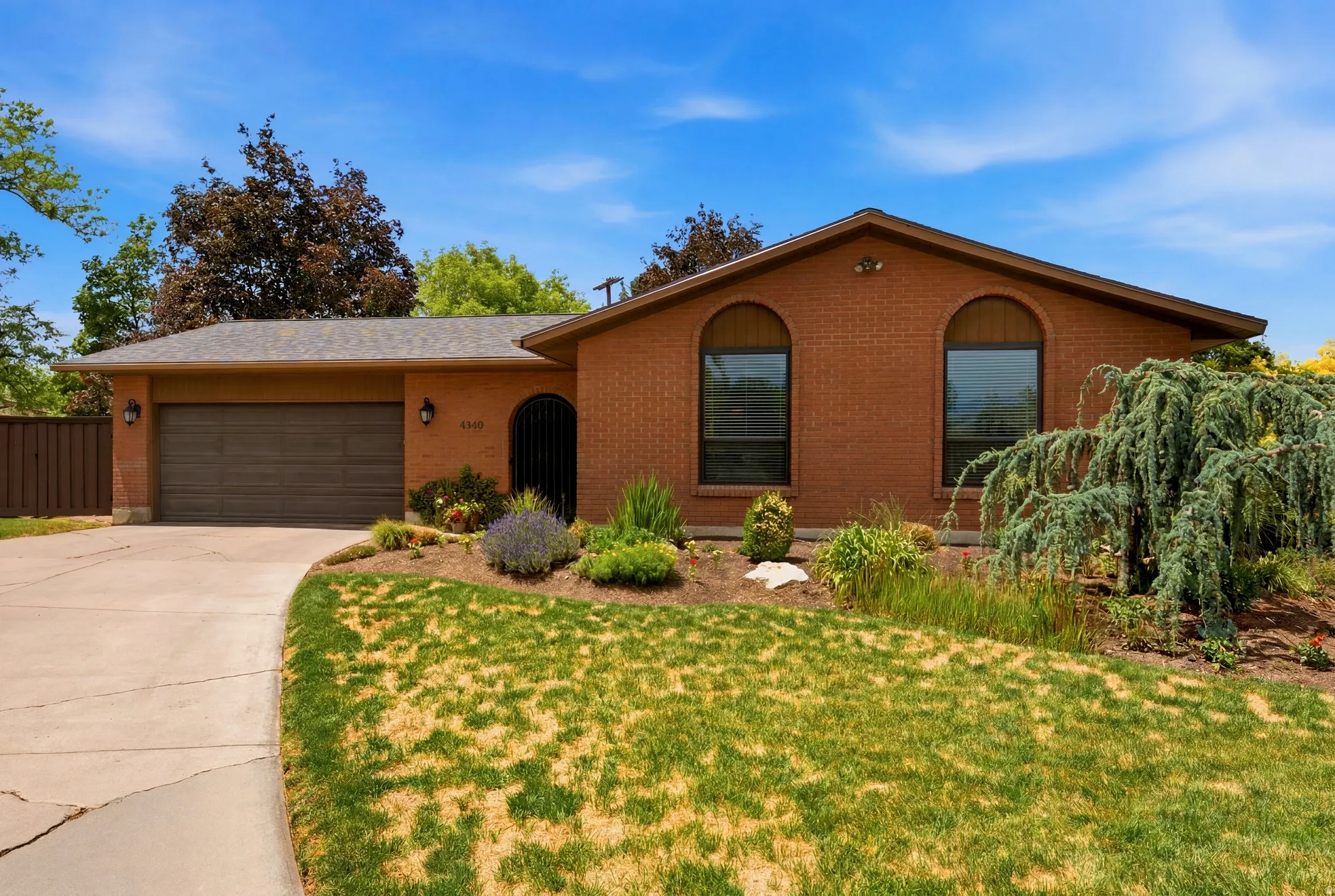 Brick exterior with arched windows, an attached garage, a paved driveway, a gabled roofline, and established front landscaping