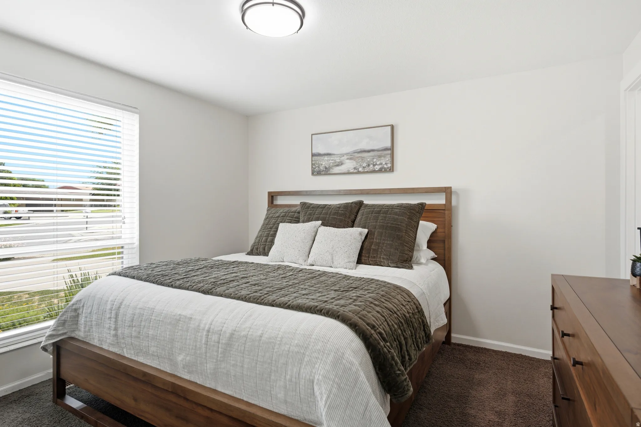 Well-lit room featuring a large window with blinds, neutral wall paint, and dark carpet flooring