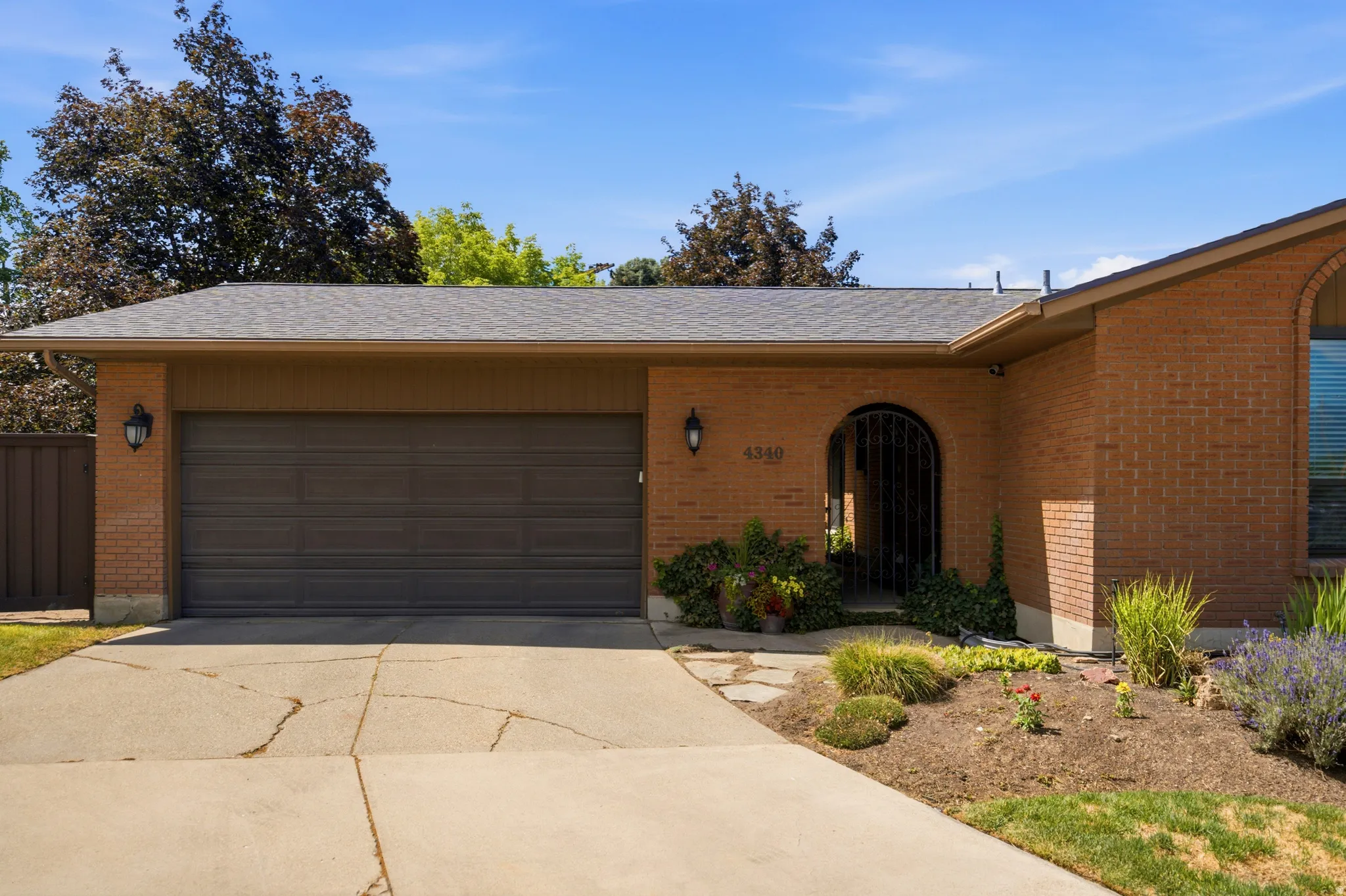 Brick exterior featuring a two-car garage with a dark brown door, an arched entryway with a decorative iron gate, sconce lighting, and a landscaped garden bed