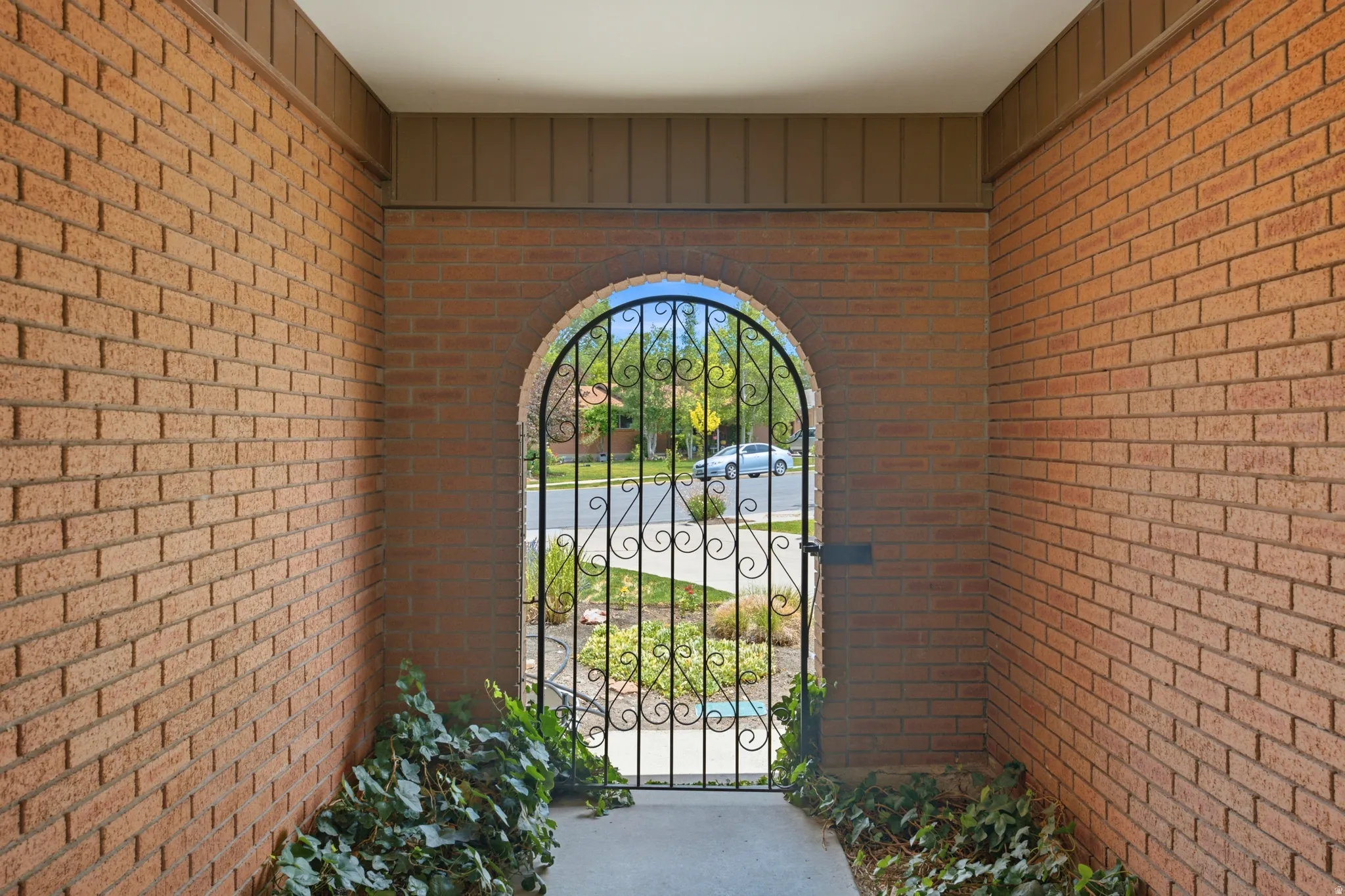 Arched wrought iron gate framed by brick walls with a concrete pathway and planted greenery