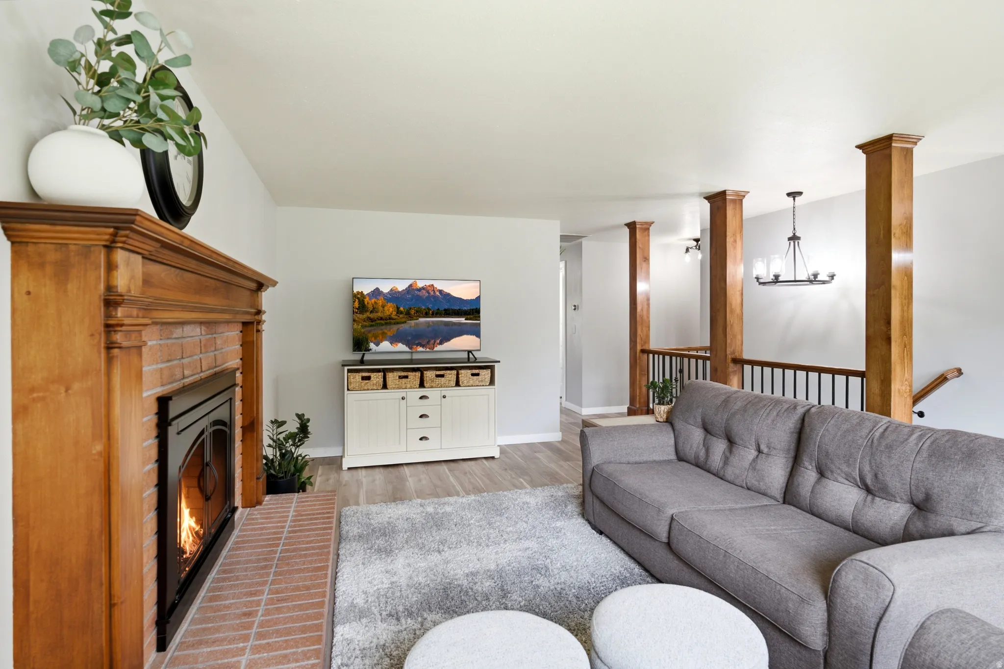 Living room featuring a brick fireplace with a wood mantle, wood-finish flooring, and wood-wrapped structural pillars
