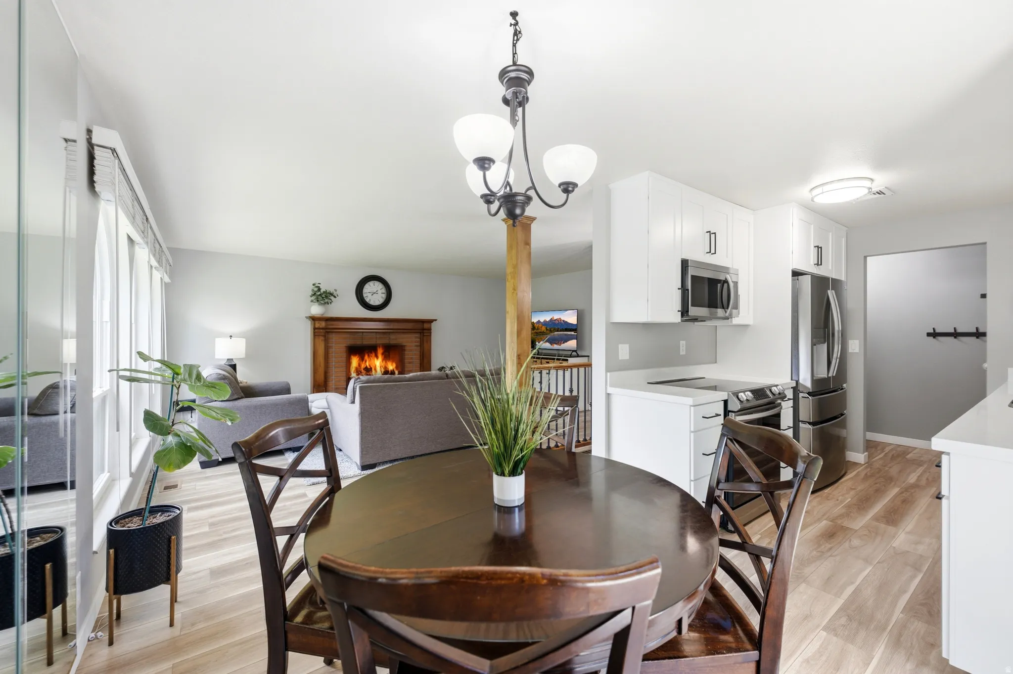 Open-concept dining area with wood-finish flooring, a contemporary chandelier, white kitchen cabinetry, stainless steel appliances, and a visible living room with a wood-mantled fireplace