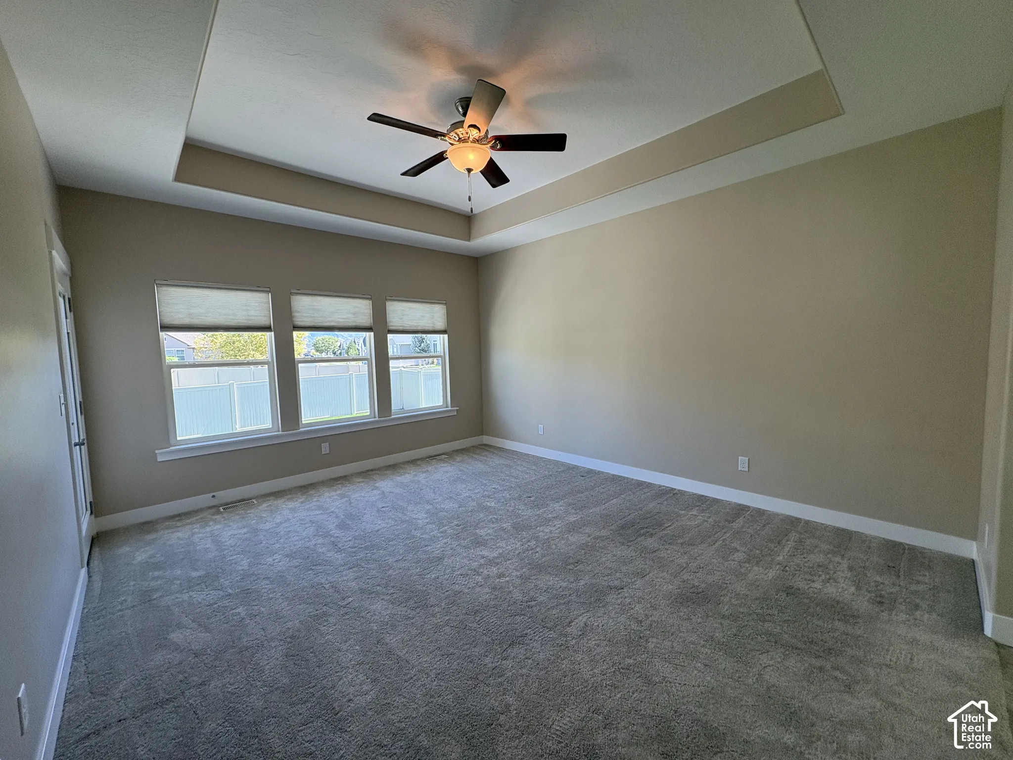 Tray ceiling with a ceiling fan, three windows, and neutral carpeting