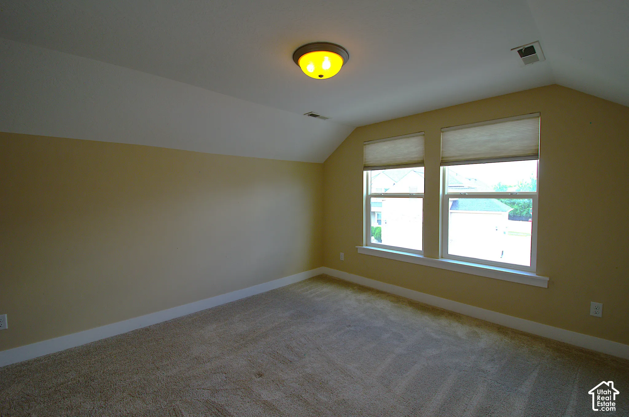 Spacious room featuring sloped ceilings, two large windows with blinds, a flush-mount ceiling light, and neutral-toned carpeting