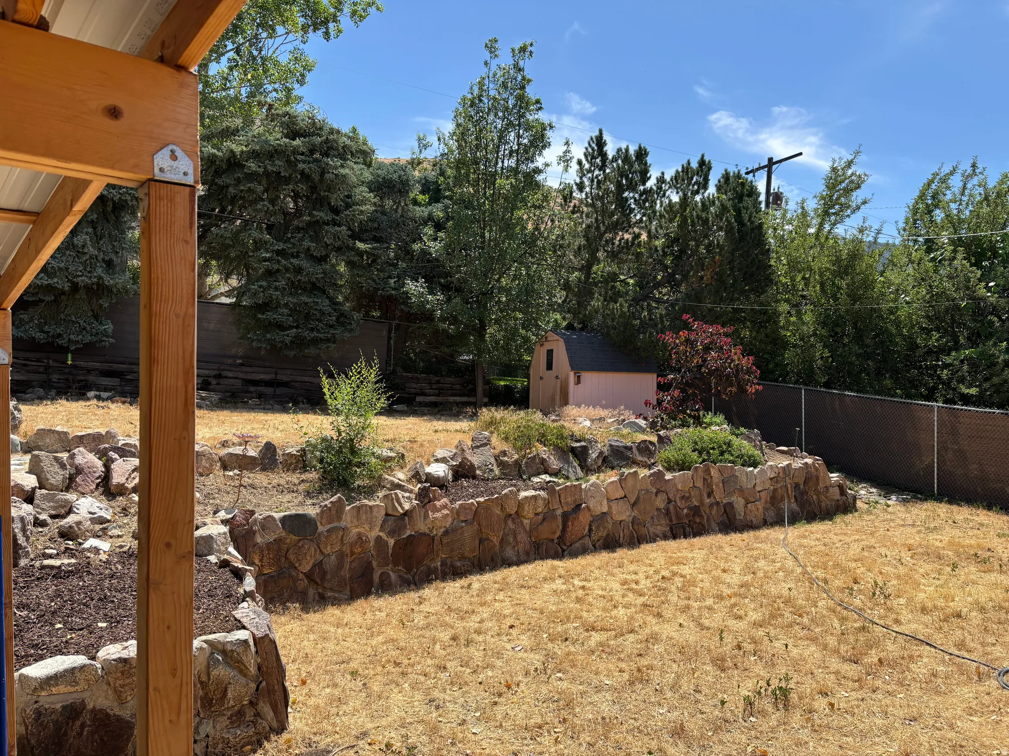 Expansive backyard featuring a stone retaining wall, varied landscaping, mature trees, and a functional shed