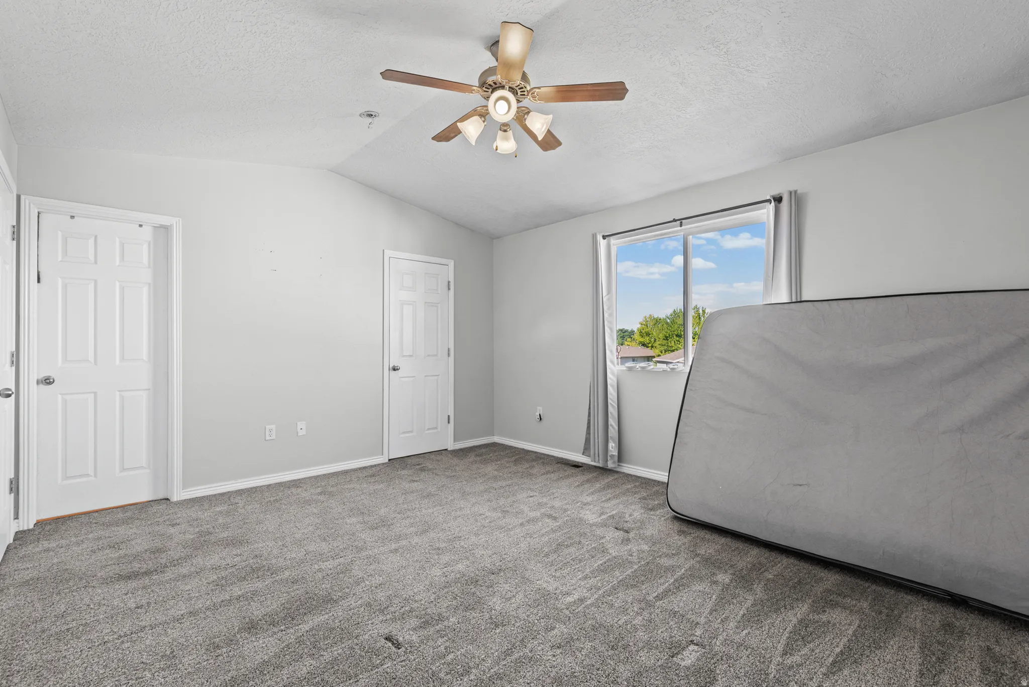 Vaulted ceiling room featuring light gray carpeting, twin pane windows with gray curtains, and a ceiling fan with light fixture