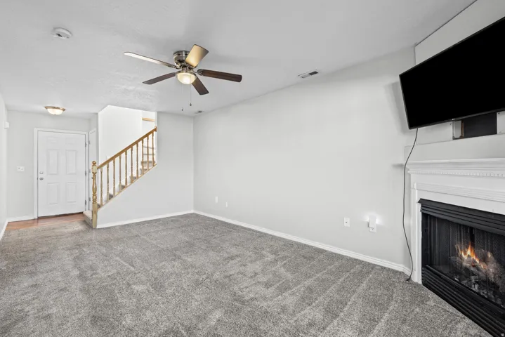 Living area featuring gray carpeting, a fireplace with a white mantel, and a ceiling fan with integrated lighting