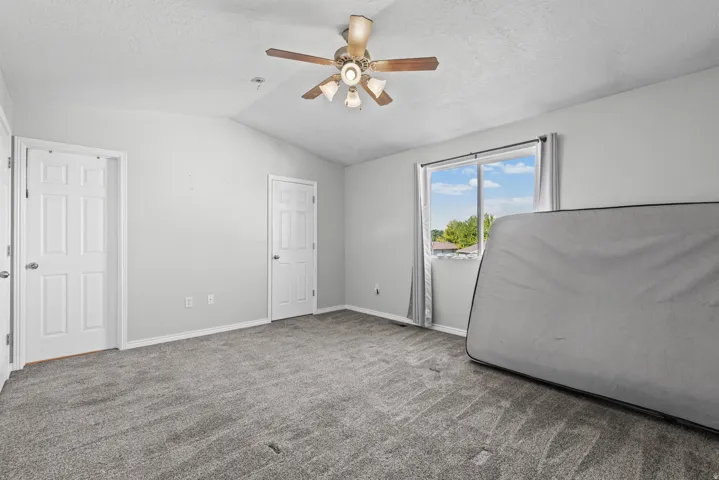 Vaulted ceiling room featuring light gray carpeting, twin pane windows with gray curtains, and a ceiling fan with light fixture