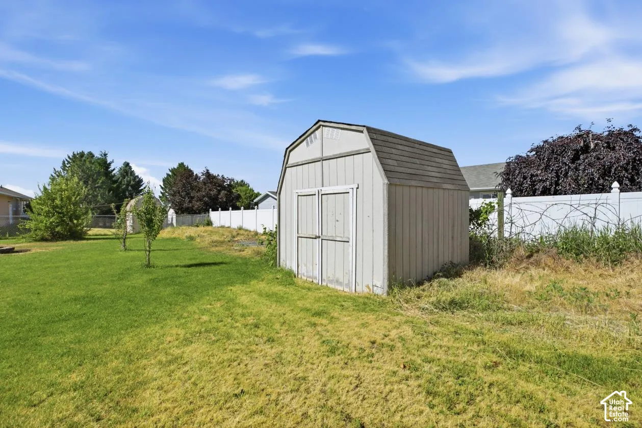 Detached storage shed with a gambrel roof and double doors, positioned within a spacious backyard featuring a green lawn and partial vinyl fencing
