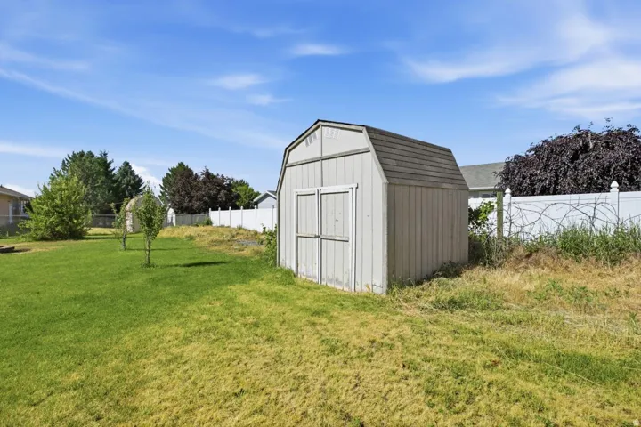 Detached storage shed with a gambrel roof and double doors, positioned within a spacious backyard featuring a green lawn and partial vinyl fencing