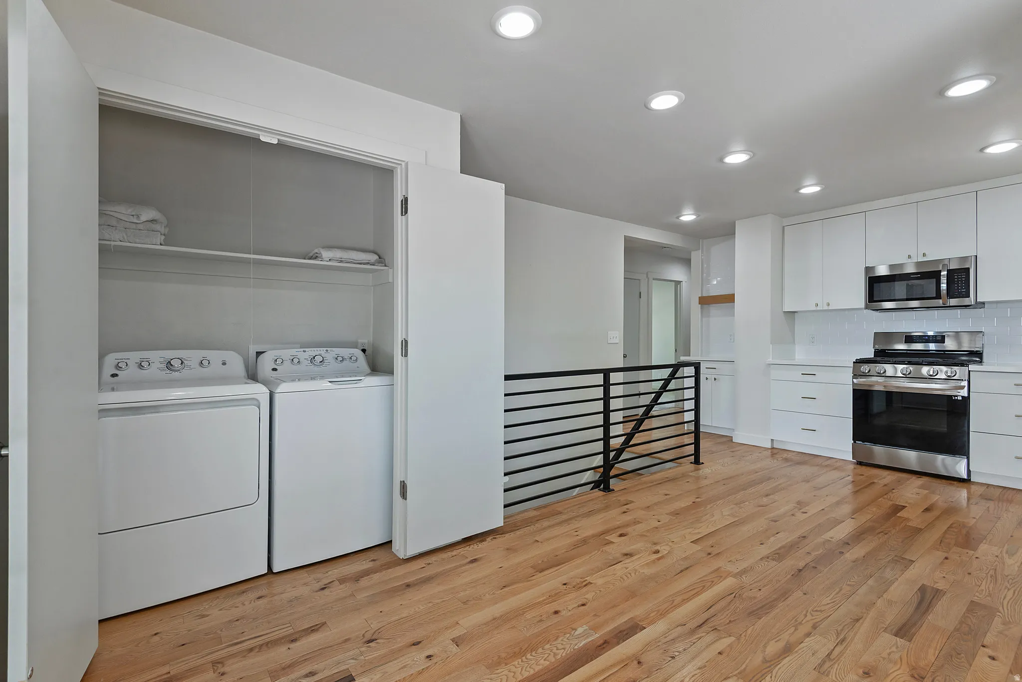 Open-concept kitchen and laundry area featuring wood-finish flooring, recessed lighting, and a modern horizontal metal railing
