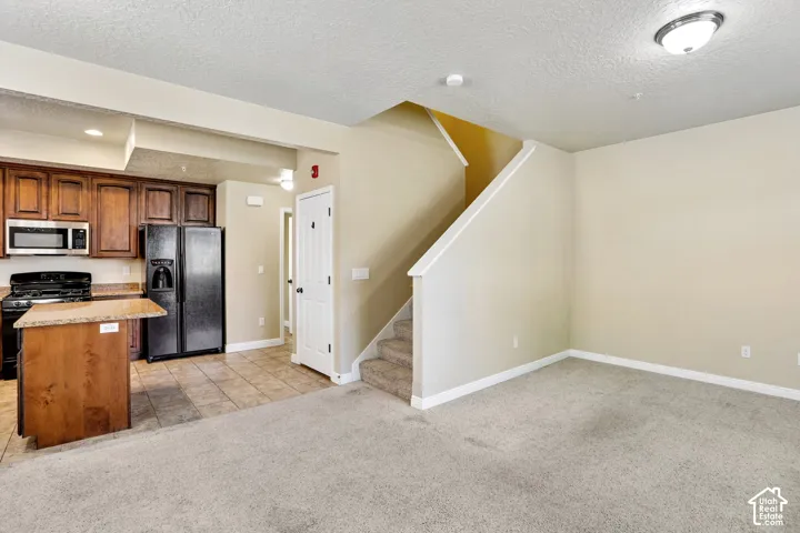Open-concept living area with new carpet, featuring a kitchen with dark wood-finish cabinetry, stainless steel microwave, black refrigerator, and a kitchen island with a stone-look countertop