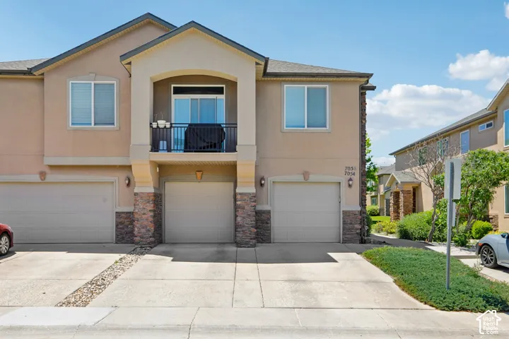 Multi-story stucco exterior featuring a prominent balcony with a black railing, three individual garage doors, stone veneer accents on the lower facade, and well-maintained landscaping