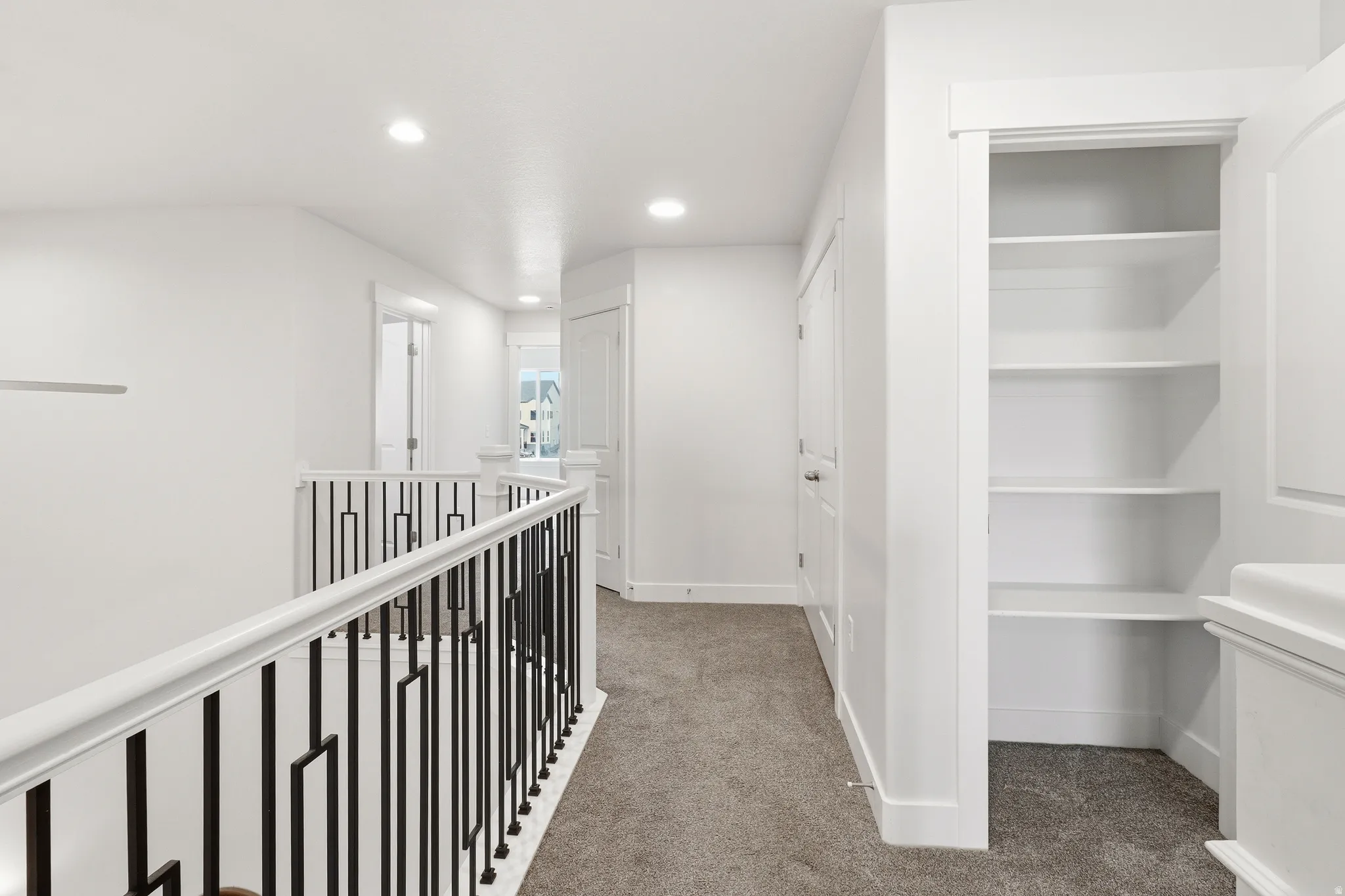 Carpeted hallway featuring a white railing with black metal spindles, recessed lighting, white walls, and built-in shelving