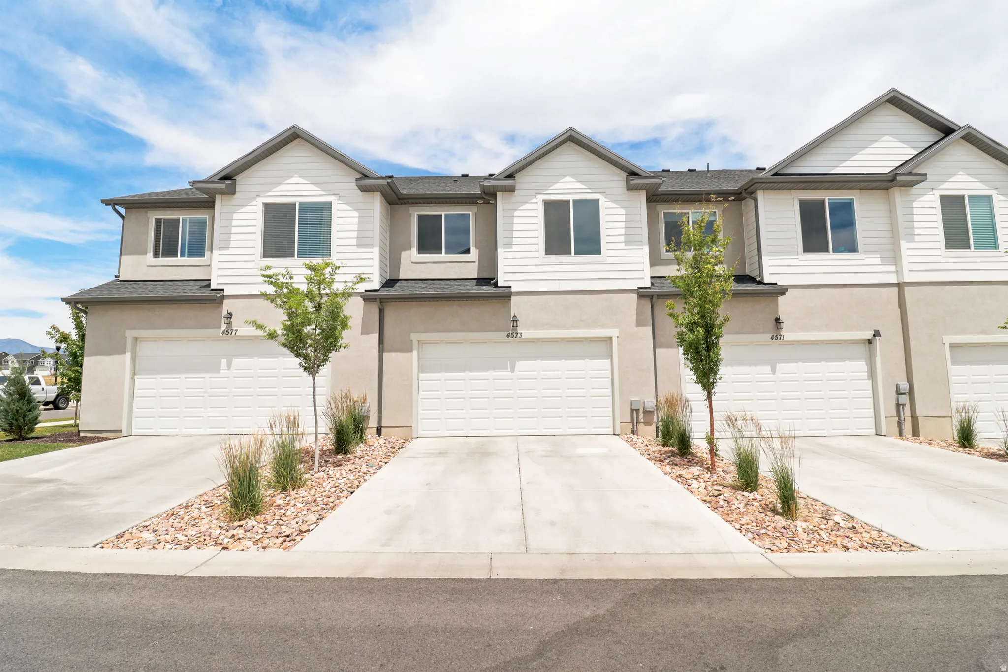 Modern townhome exterior featuring light-colored siding, a two-car garage, established landscaping, and a concrete driveway