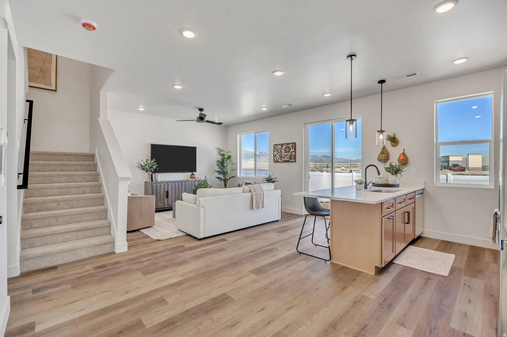Open-concept living space featuring wood-finish flooring, recessed lighting, a kitchen island with seating, a stainless steel sink with a black faucet, and large windows