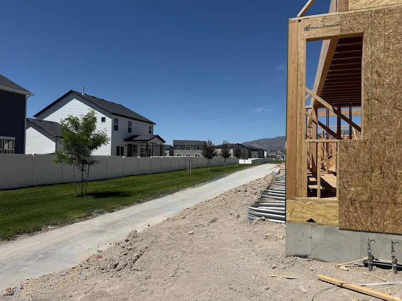 New construction framing with visible foundation, surrounded by established homes featuring white siding and dark roofing