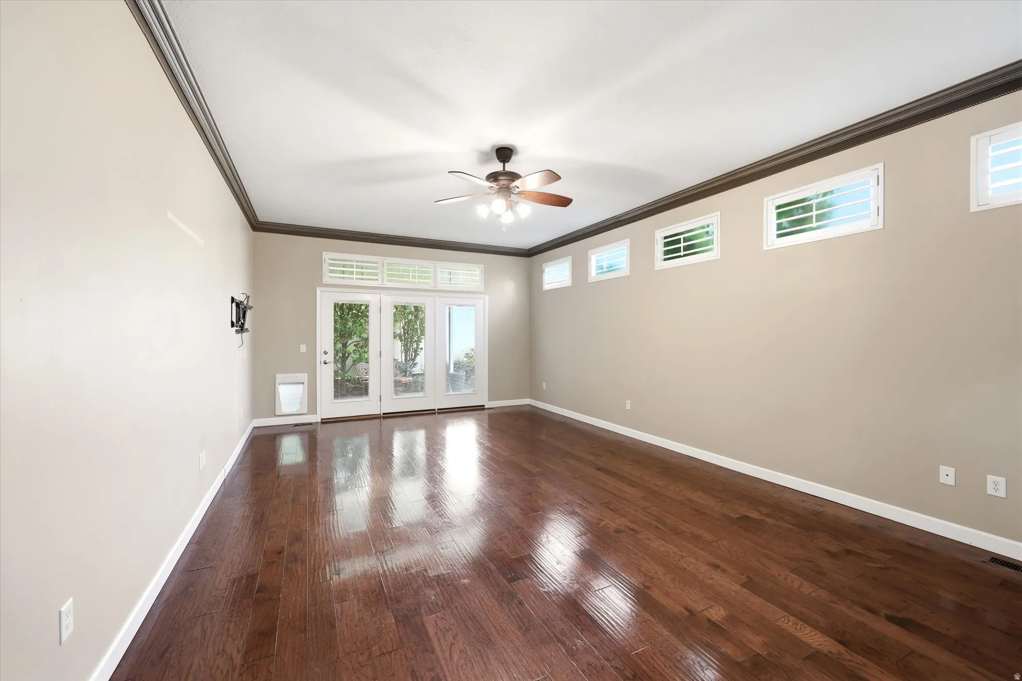 Spacious Main bedroom featuring rich wood-finish flooring, a ceiling fan with integrated lighting, and multiple transom windows with French doors to access the backyard.
