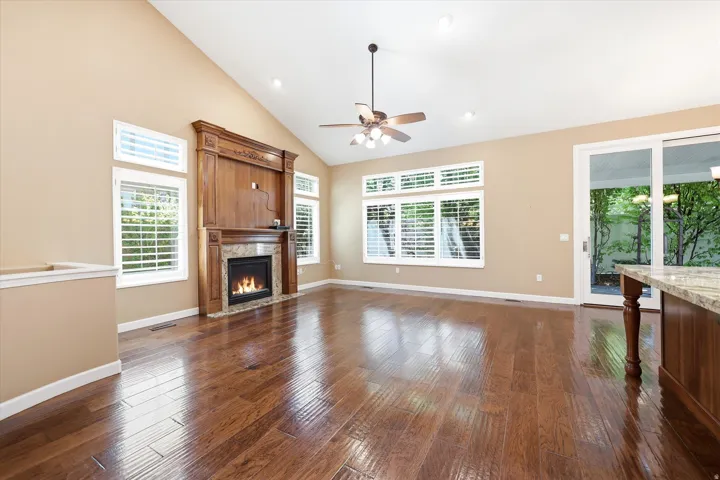 Great room featuring vaulted ceilings, an ornate wood-mantled fireplace, lustrous wood-finish flooring, a ceiling fan with integrated lighting, and multiple windows with plantation shutters