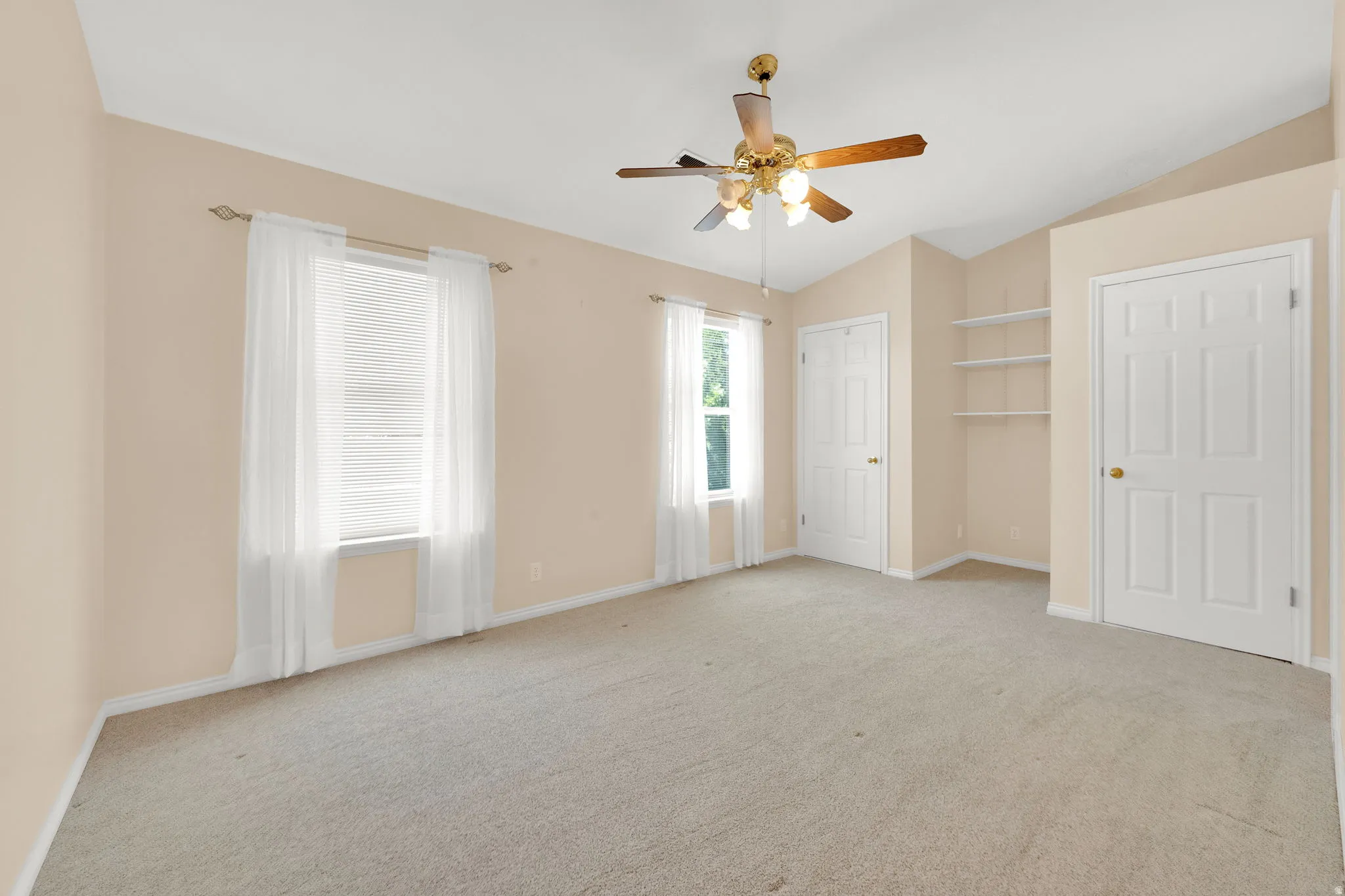 Spacious room featuring vaulted ceilings, a ceiling fan with light fixture, light-colored carpet flooring, two windows with blinds and sheer curtains, and built-in shelving