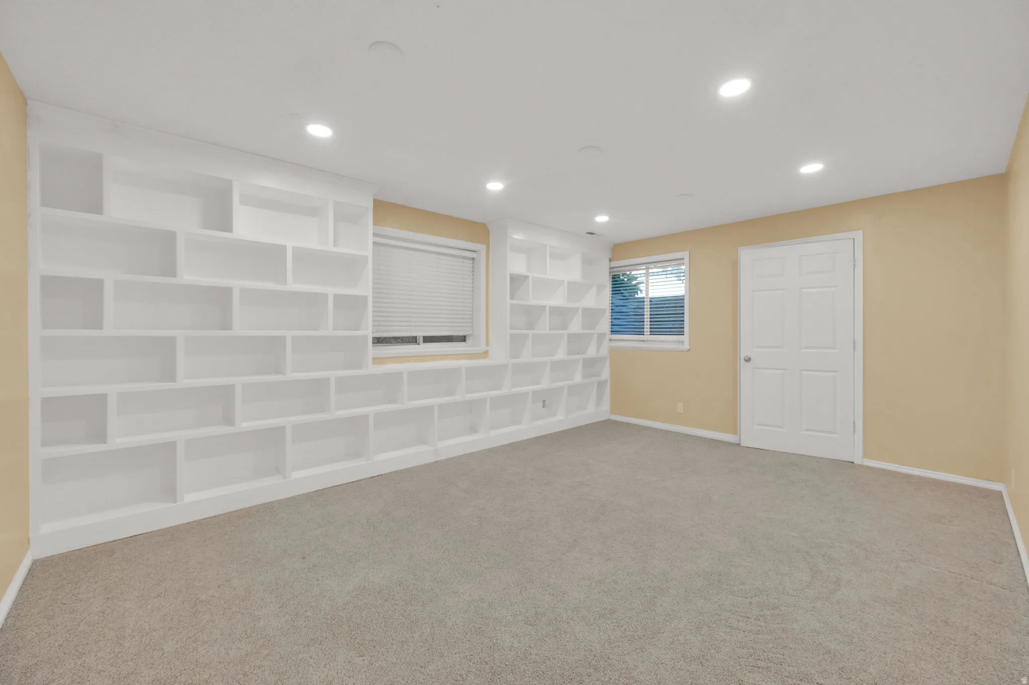 Expansive room featuring a full wall of white built-in shelving, two windows with blinds, recessed lighting, and light-colored carpeting